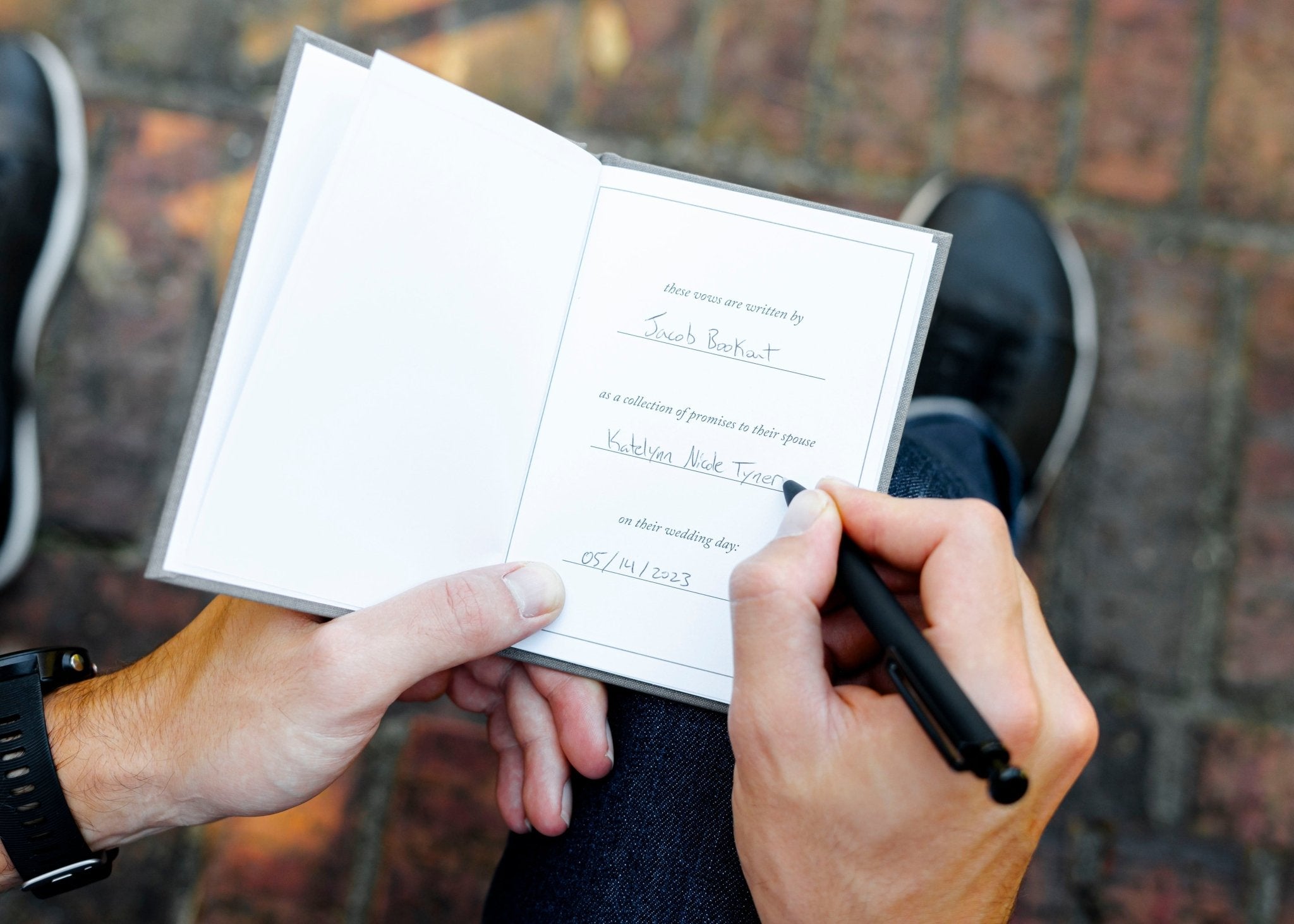 Hands holding open white vow book with handwritten text inside, displayed outdoors against brick background.