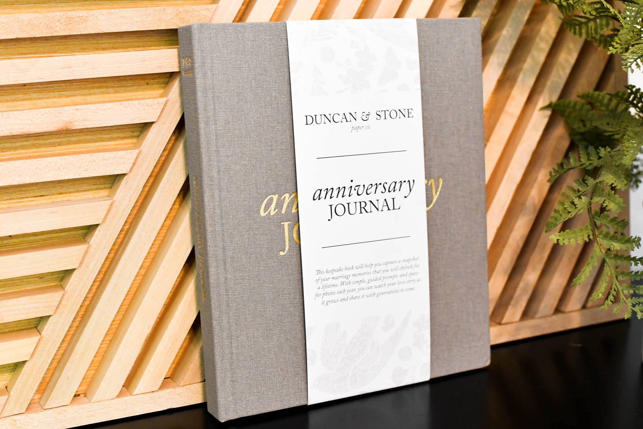 Gray linen anniversary journal with white belly band labeled "Duncan & Stone Anniversary Journal" on wooden striped background.