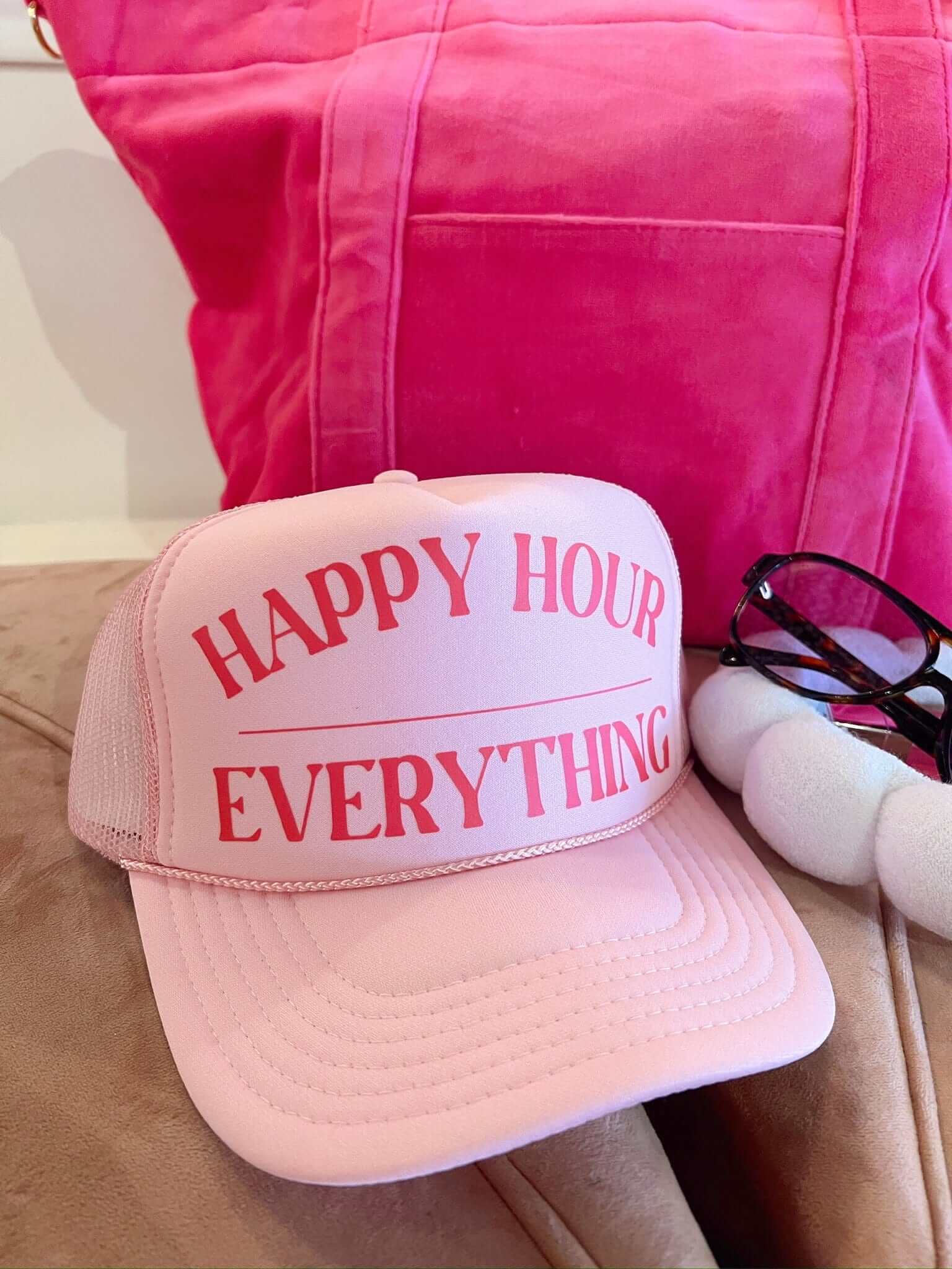 Light pink trucker hat with red "HAPPY HOUR EVERYTHING" text, displayed with pink tote bag and sunglasses.