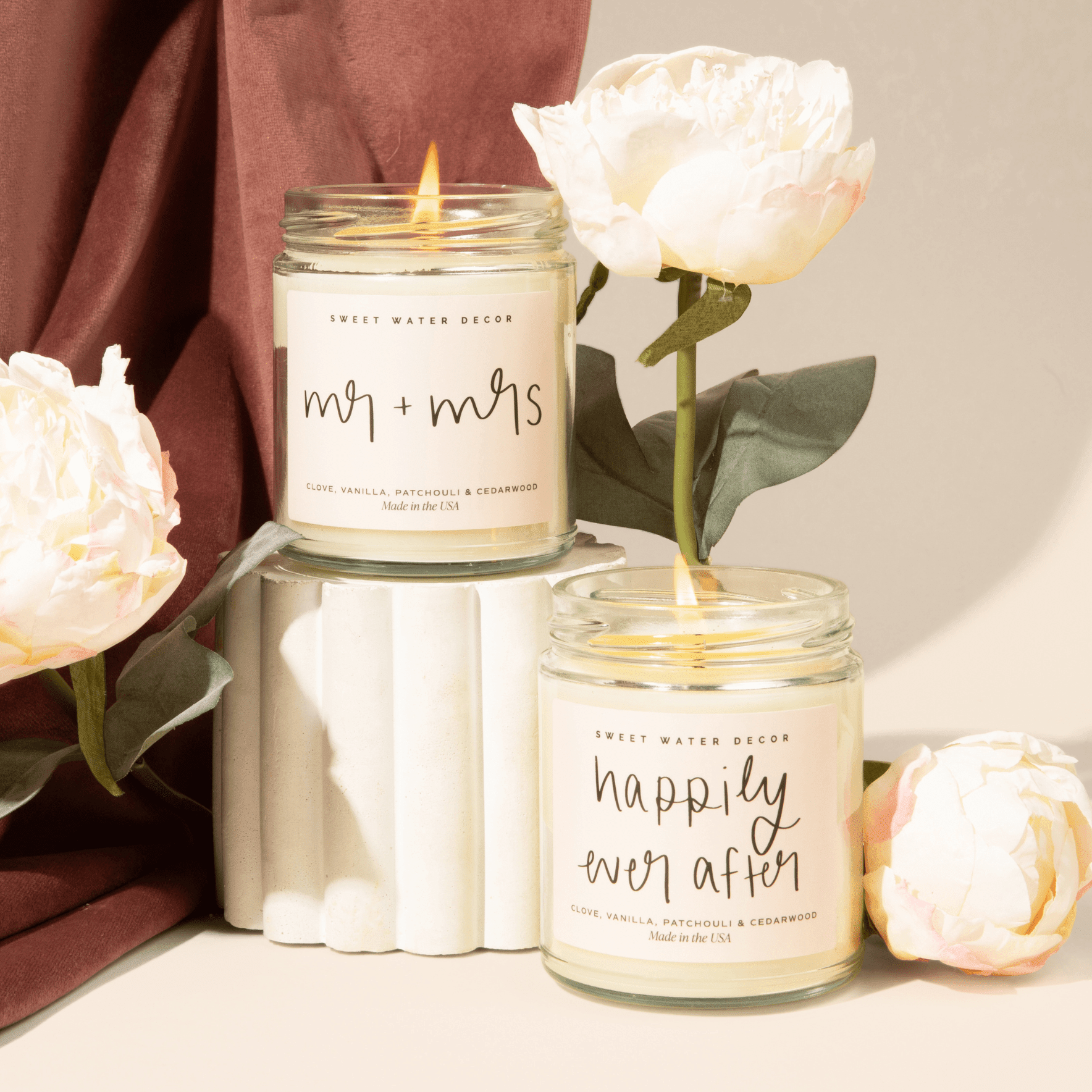 Two cream soy candles with "mr + mrs" and "happily ever after" text on glass jars, surrounded by white roses and burgundy fabric.
