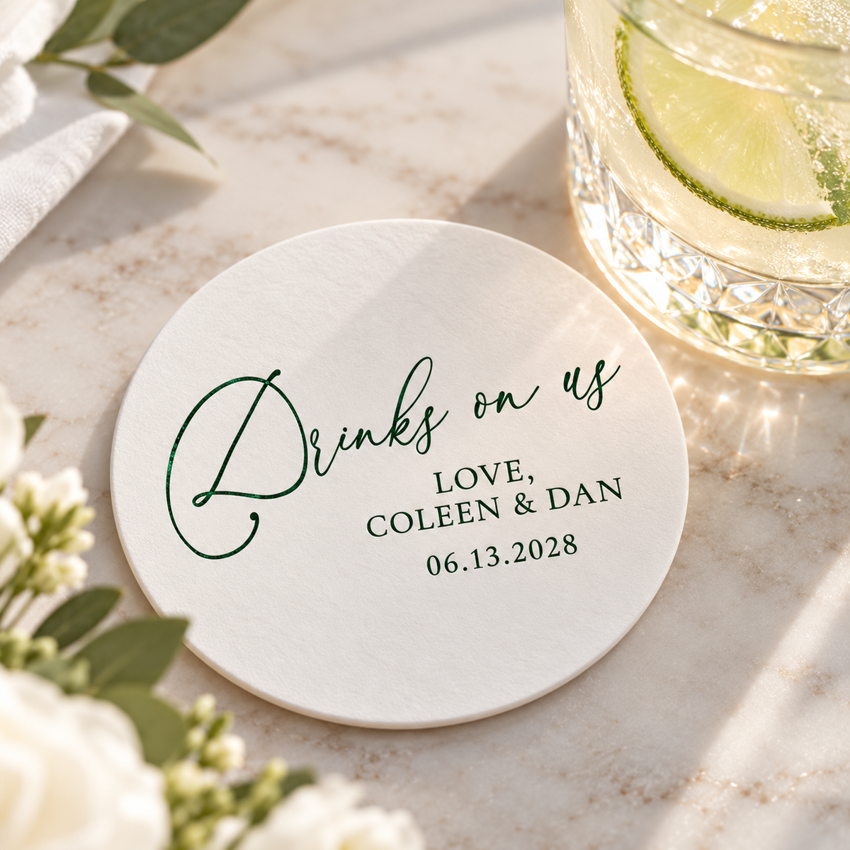Custom Drinks on Us wedding coaster with dark green foil lettering, couple names and wedding date, styled beside a lime cocktail on a marble table.