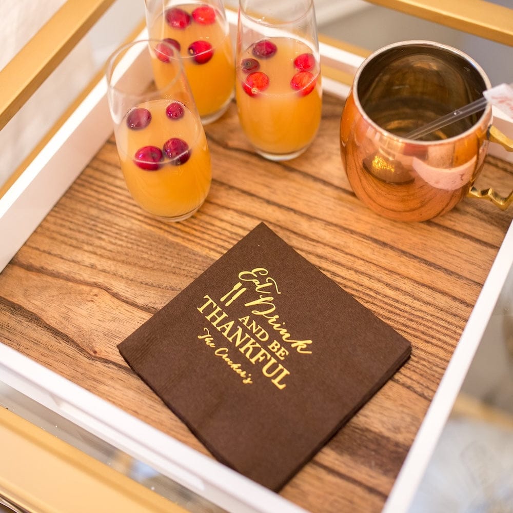Black foil-stamped napkins with "Eat Drink and Be Thankful" design displayed on wooden tray with orange cocktails and copper mug.