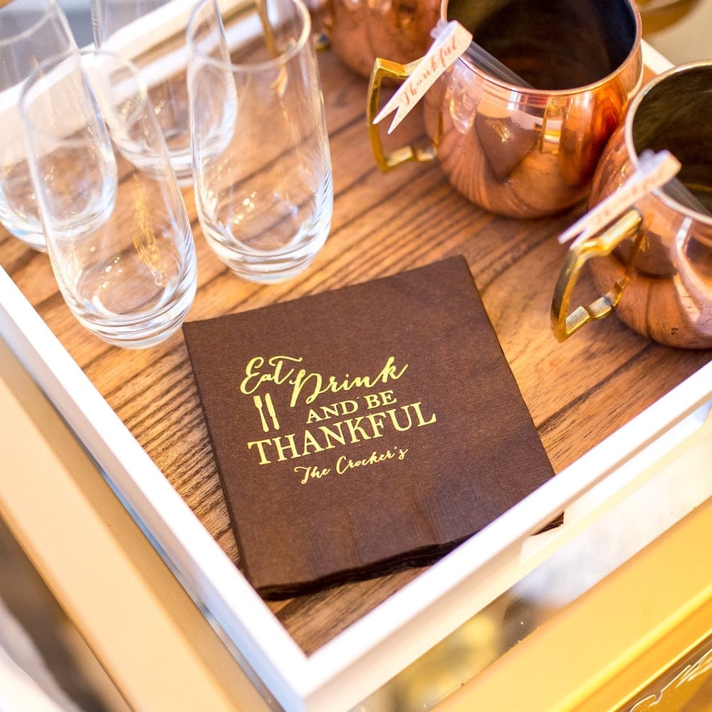 Brown foil-stamped napkin with "Eat Drink and Be Thankful" text and personalization, displayed on wooden tray with glasses and copper pitchers.