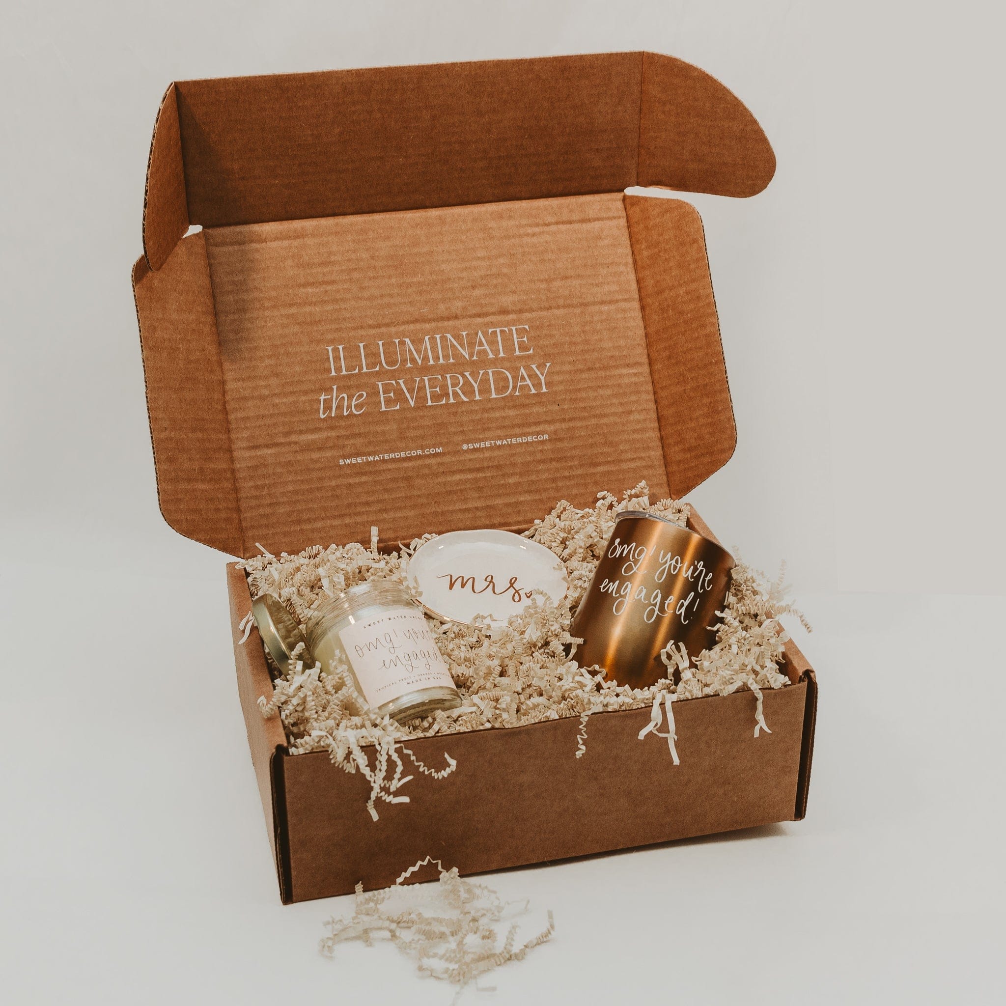 Open kraft gift box with "Illuminate the Everyday" engraved on lid, containing gold candle, matches, and white card nestled in straw filler.