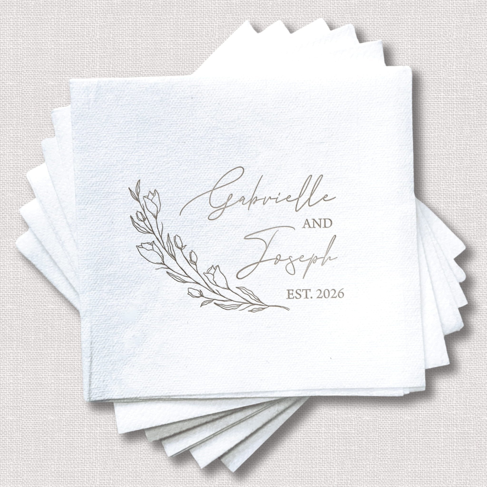 Stack of white linen napkins with gray foil-stamped names "Gabrielle and Joseph" and botanical leaf design, EST. 2026