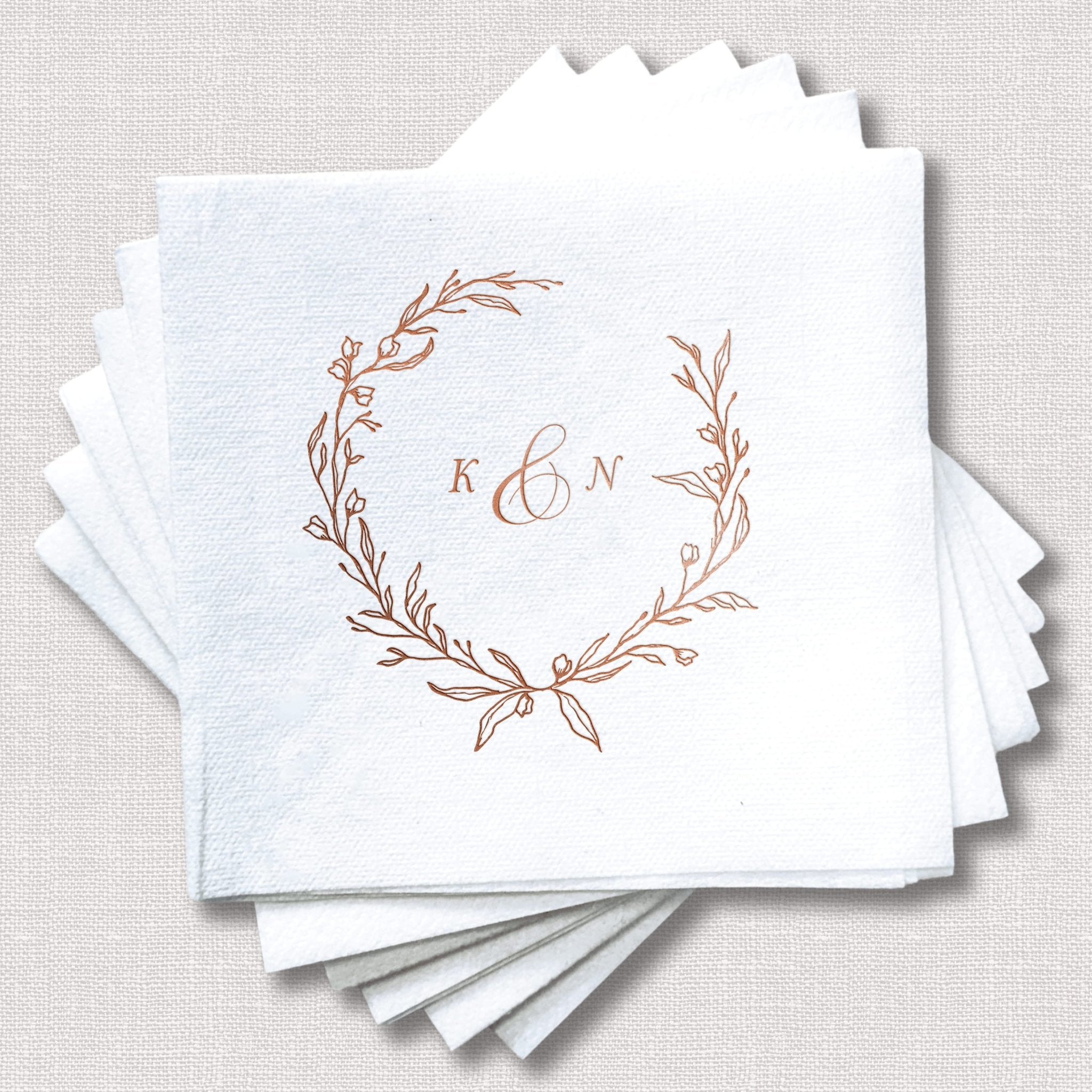Stack of white cocktail napkins with rose gold foil wreath design and personalized monogram initials K and N in center.