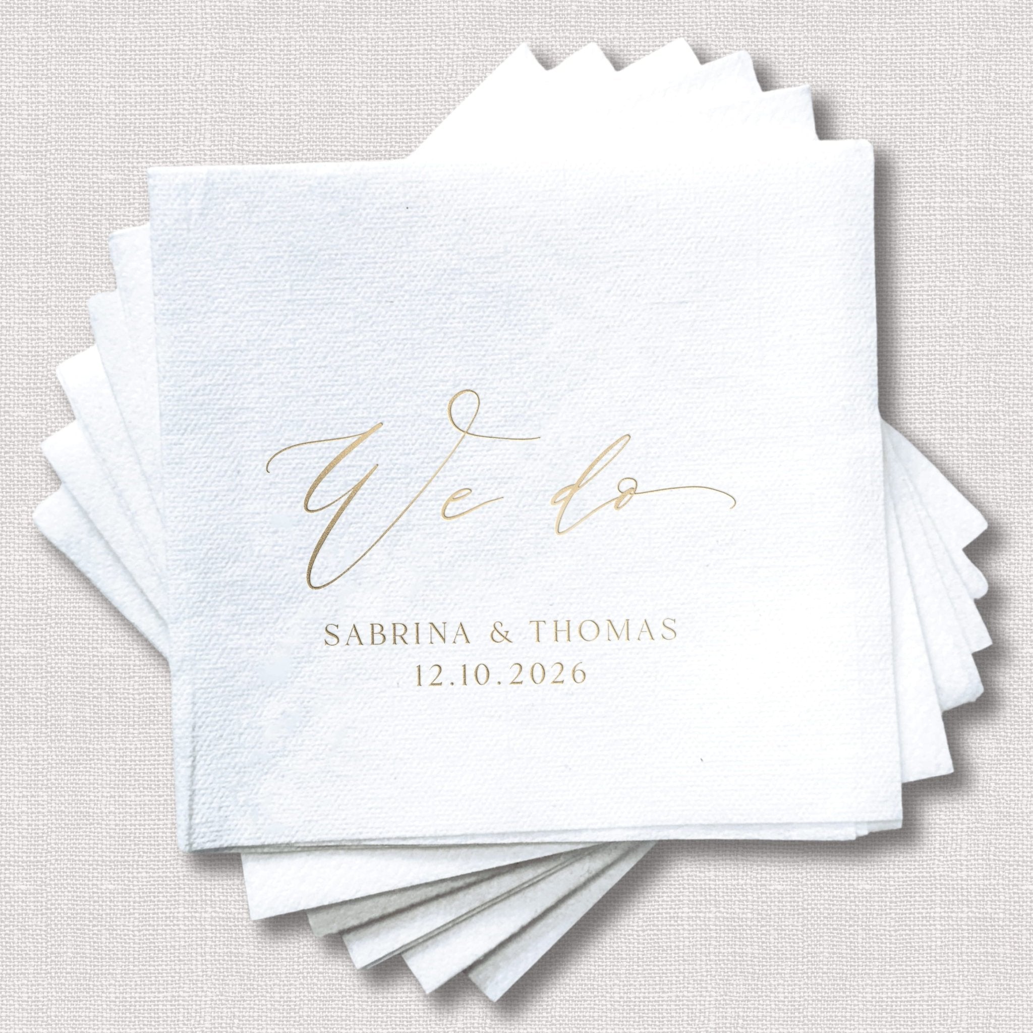 Stack of white linen napkins with gold foil "We do" script and couple names "Sabrina & Thomas" with wedding date 12.10.2026