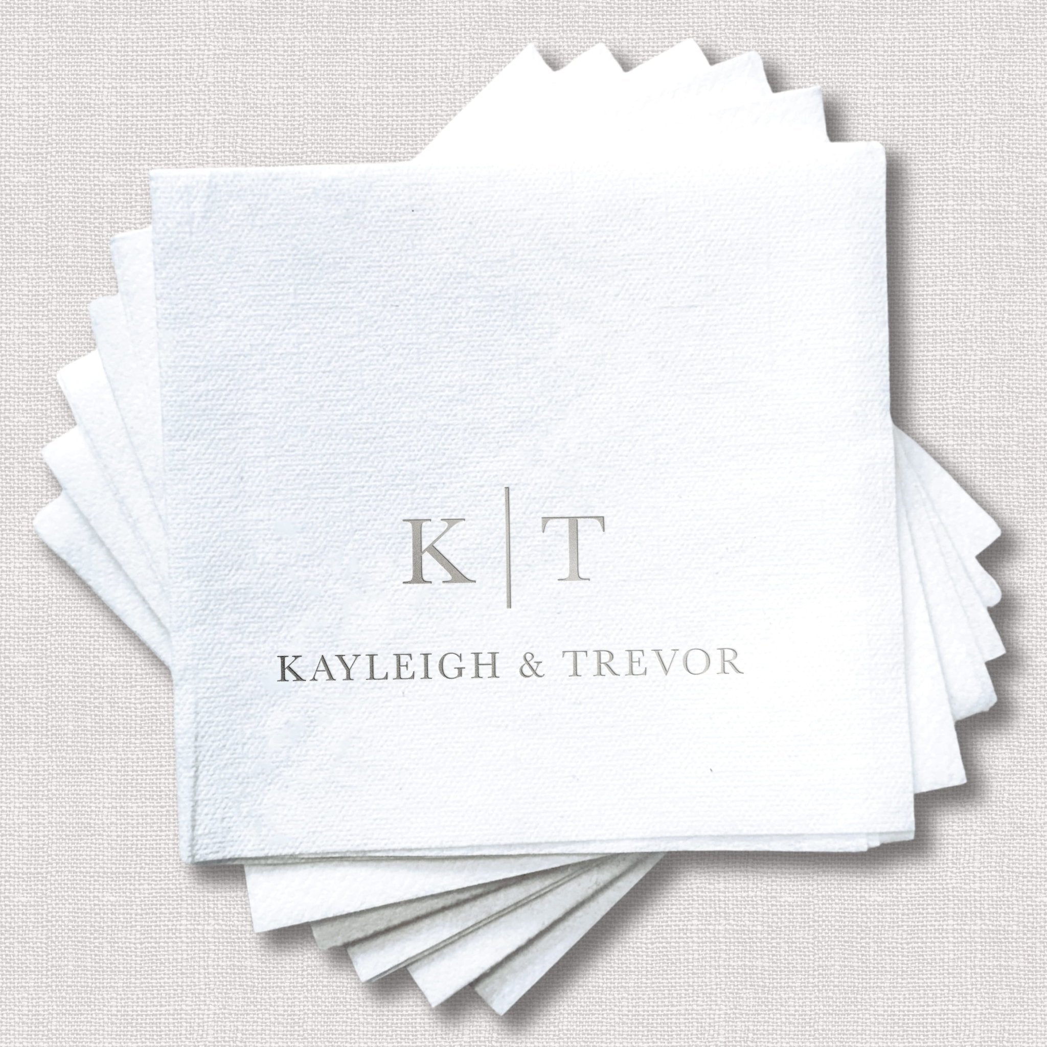 Stack of white linen napkins with gray foil-stamped initials K|T and names Kayleigh & Trevor for a wedding.