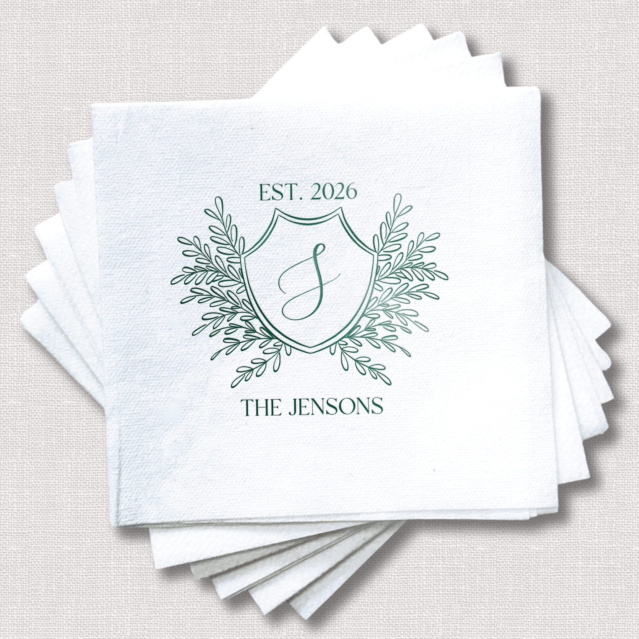 Stack of white linen napkins with green foil-stamped crest design featuring "EST. 2026," decorative leaf flourishes, initial "J," and "THE JENSONS" text.