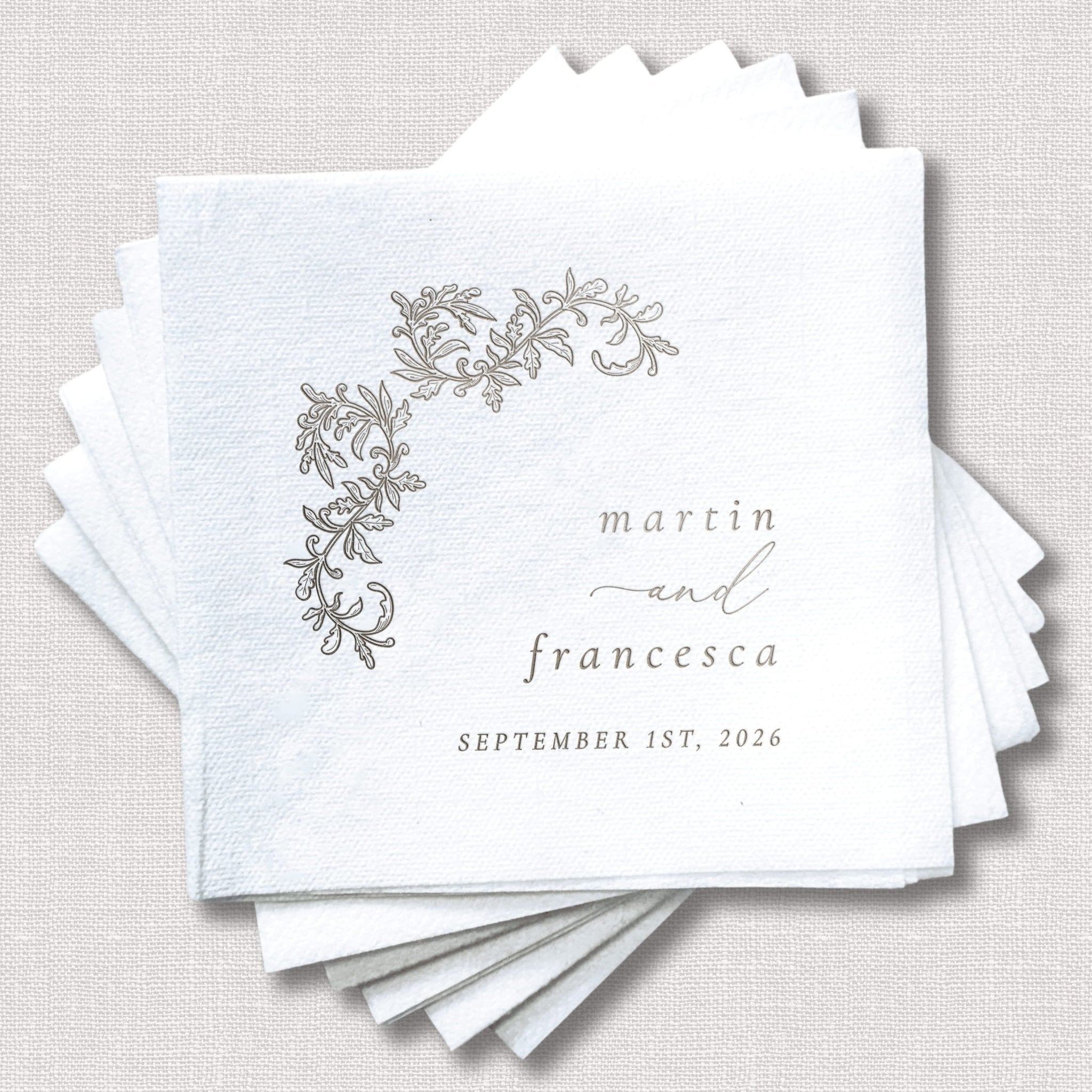 Stack of white cocktail napkins with gray floral crest design and personalized text reading "marrin and francesca september 1st, 2026"