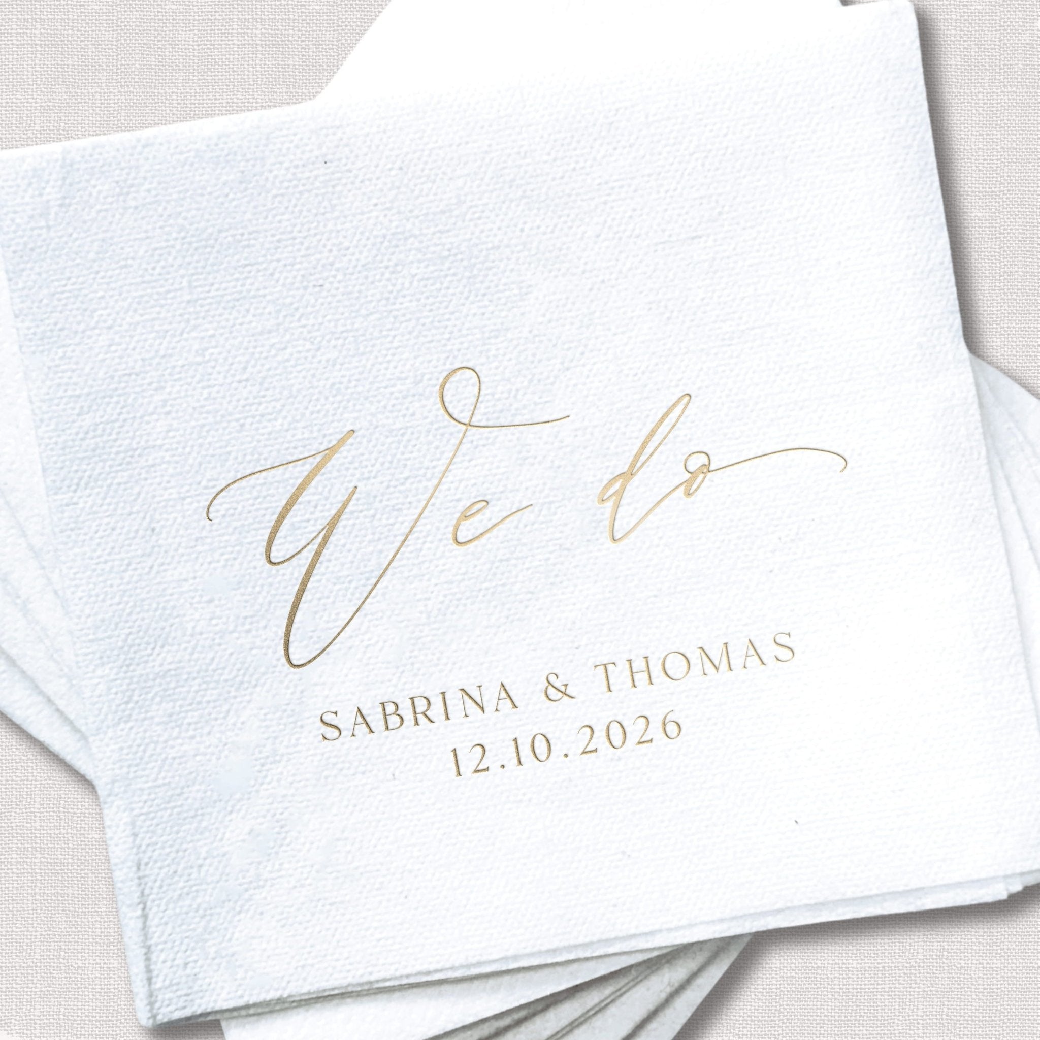 White linen napkin with gold foil stamped "we do" script and couple names "Sabrina & Thomas" with wedding date 12.10.2026