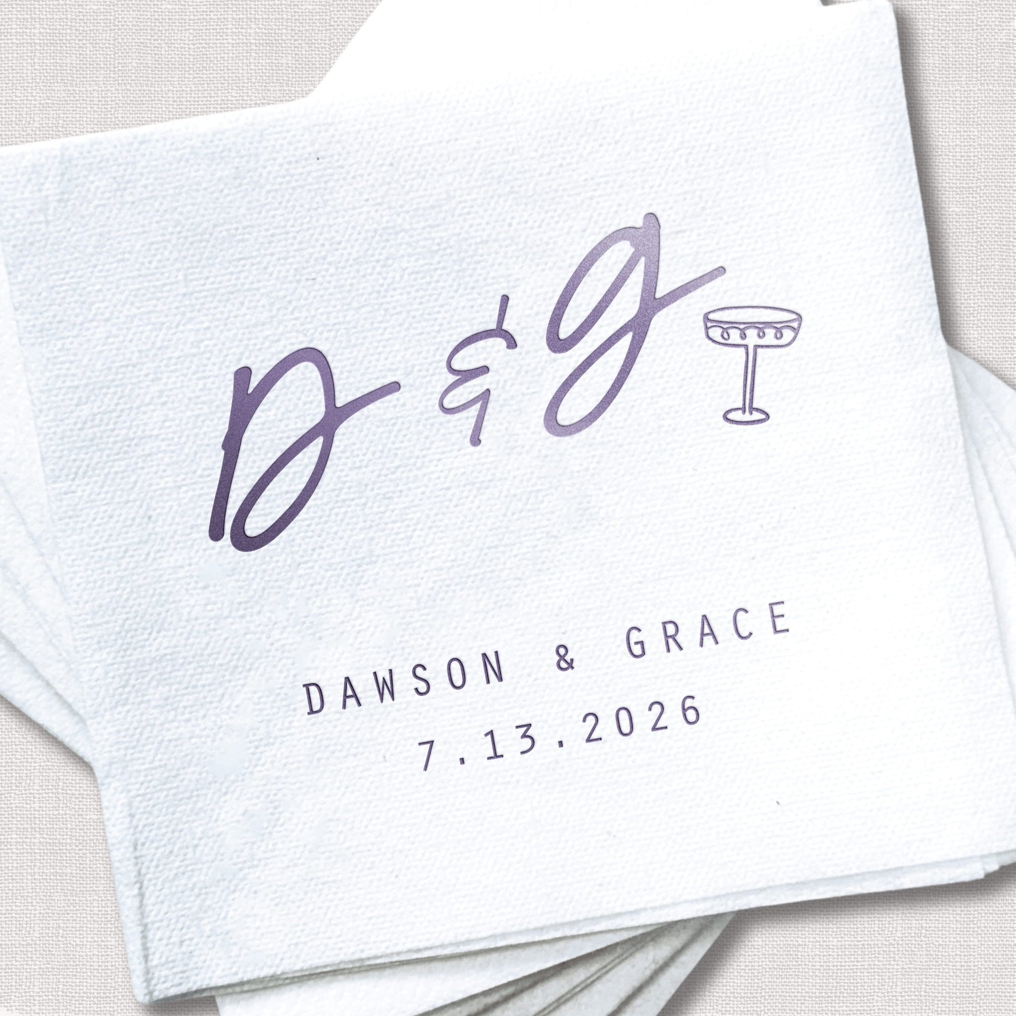 White cocktail napkins with purple foil stamped initials D & G and champagne glass, personalized with names Dawson & Grace and wedding date 7.13.2026