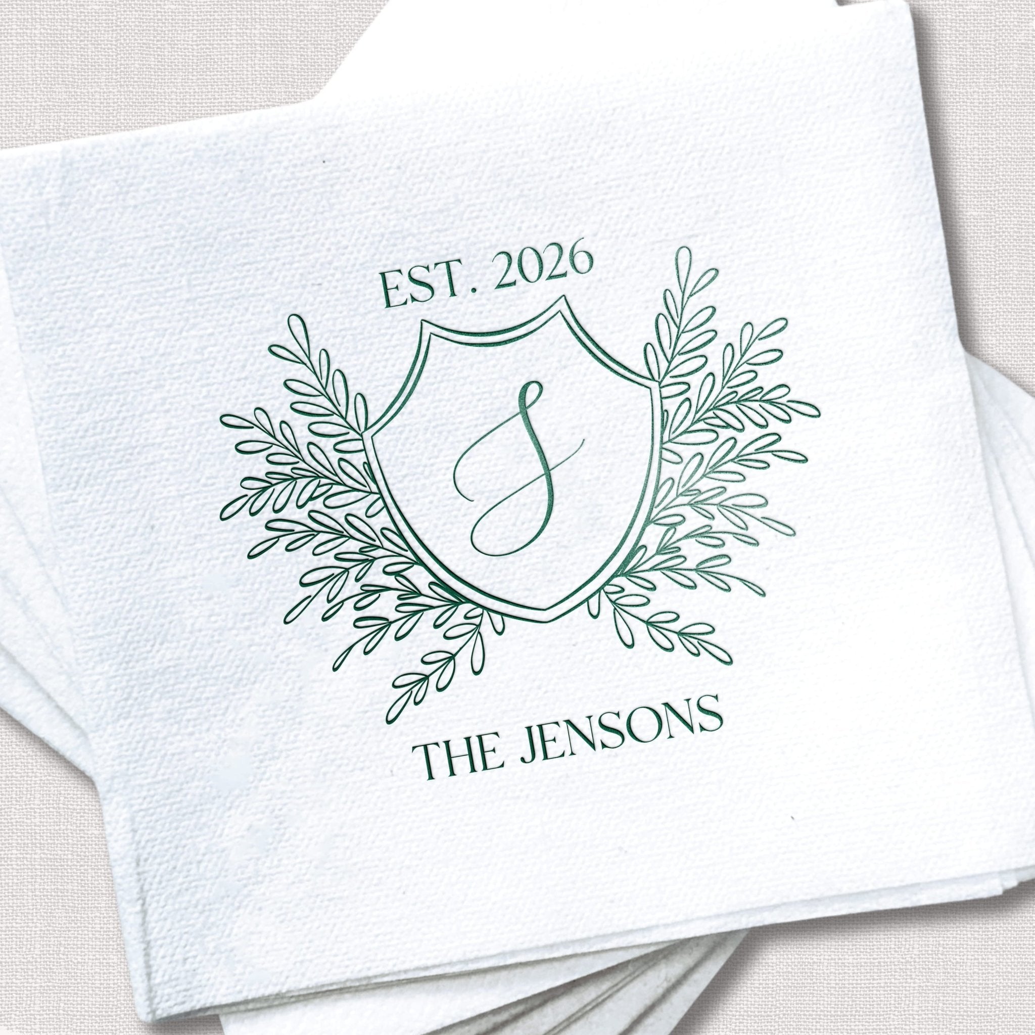 White linen napkin with green foil-stamped crest design featuring "EST. 2026," ornamental leaf flourishes, decorative monogram "J," and "THE JENSONS" text.