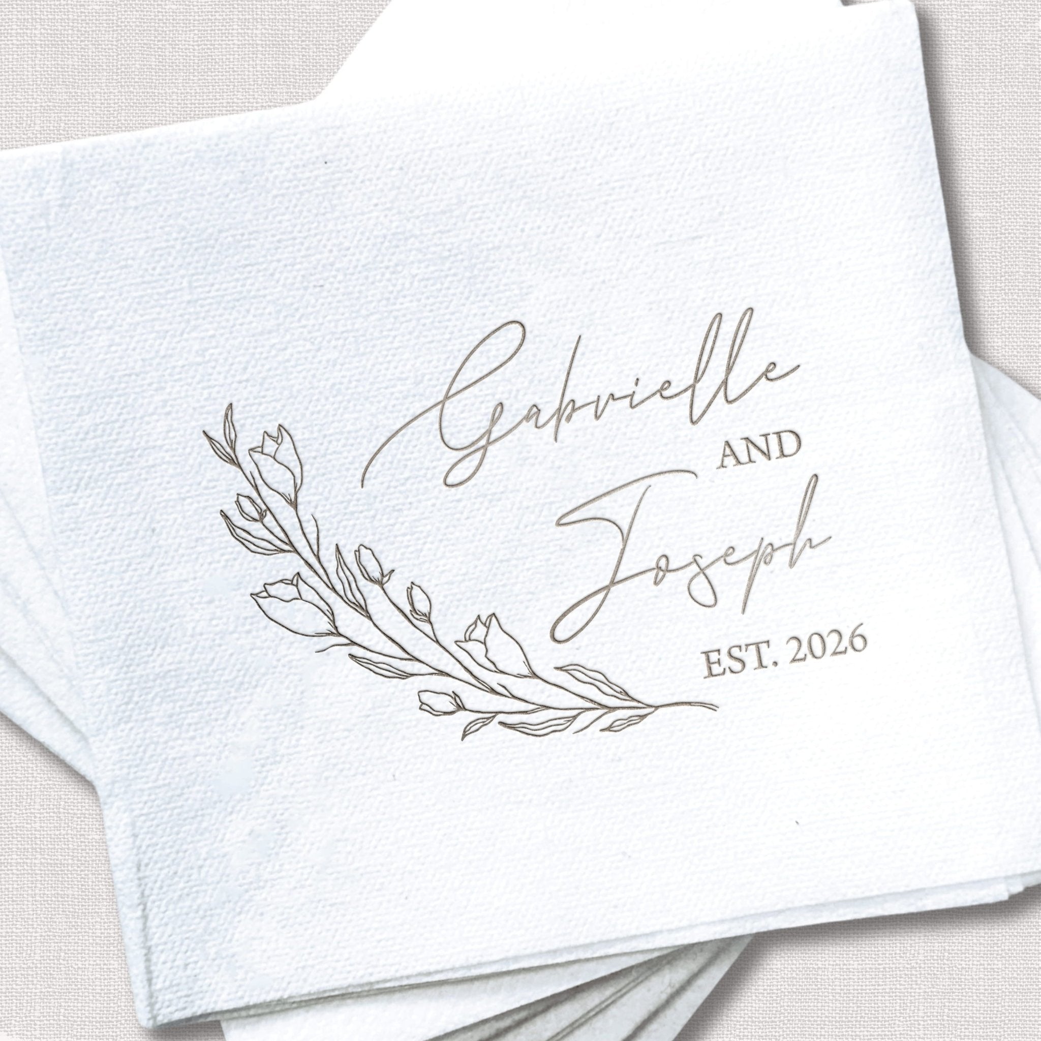 White linen napkin with gray foil-stamped names "Gabrielle and Joseph" and "EST. 2026" with delicate leaf design