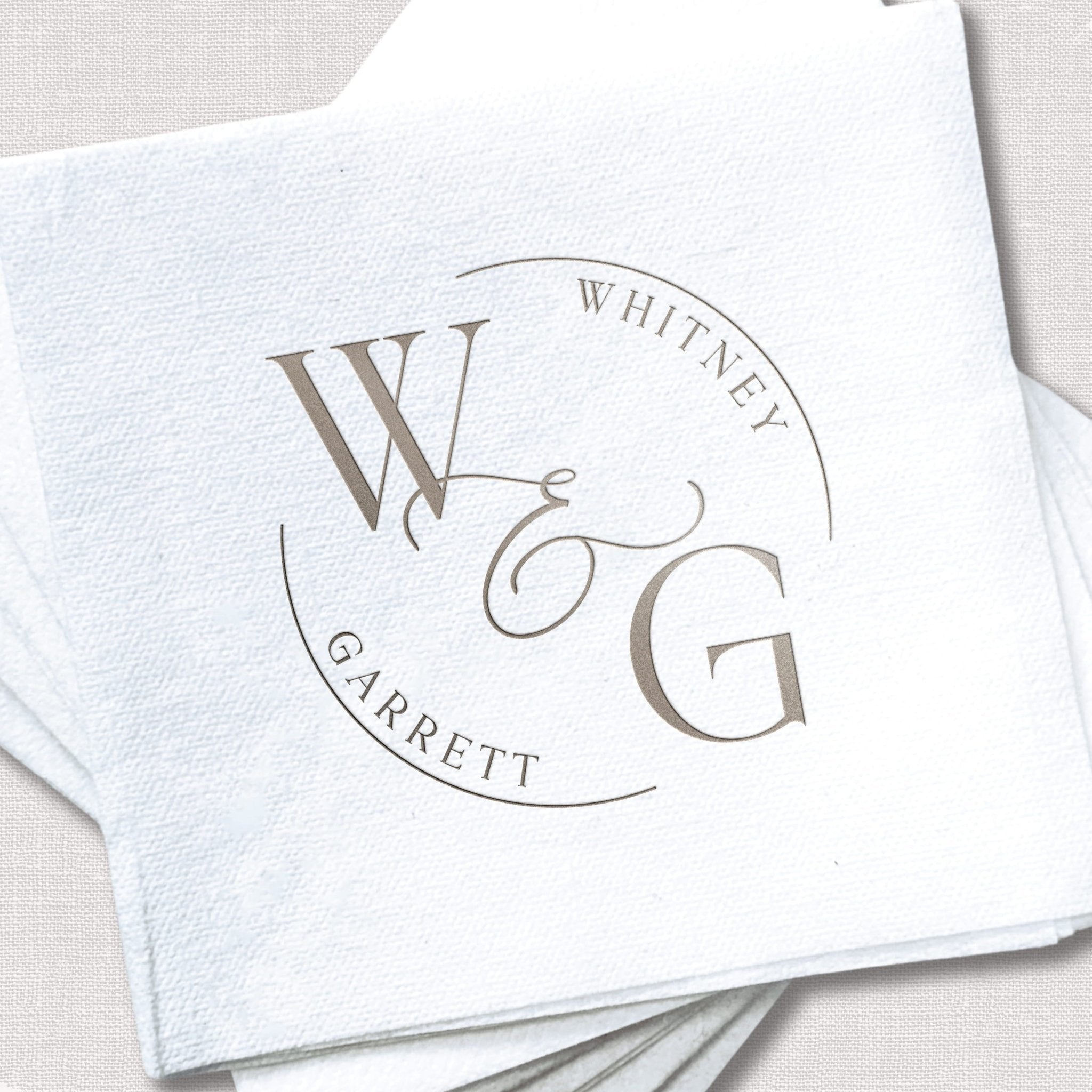 White linen cocktail napkins with silver foil monogram featuring initials W, G, and names Whitney and Garrett in circular design.