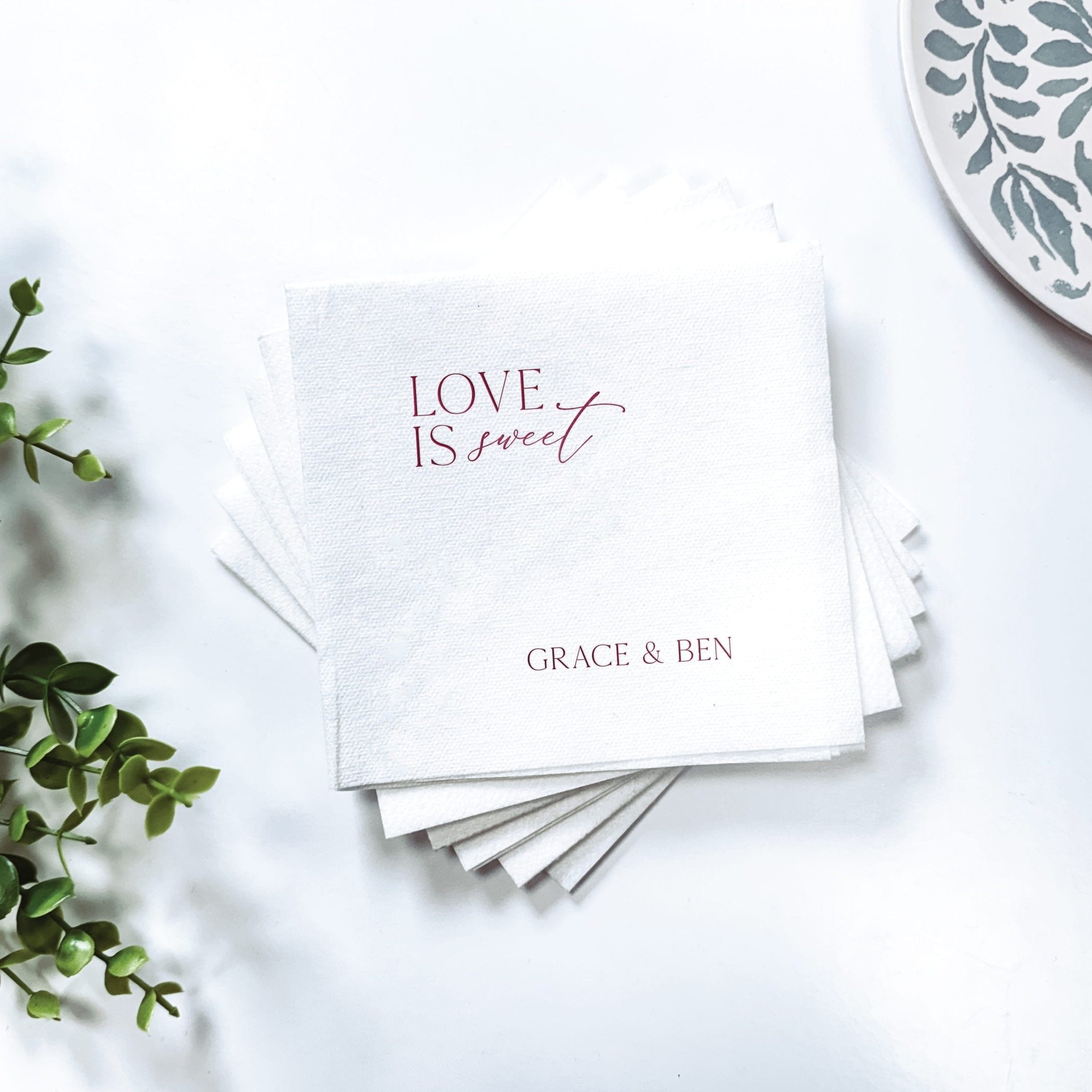 Stack of white linen napkins with "LOVE IS SWEET" text and personalized names "GRACE & BEN" in foil stamping