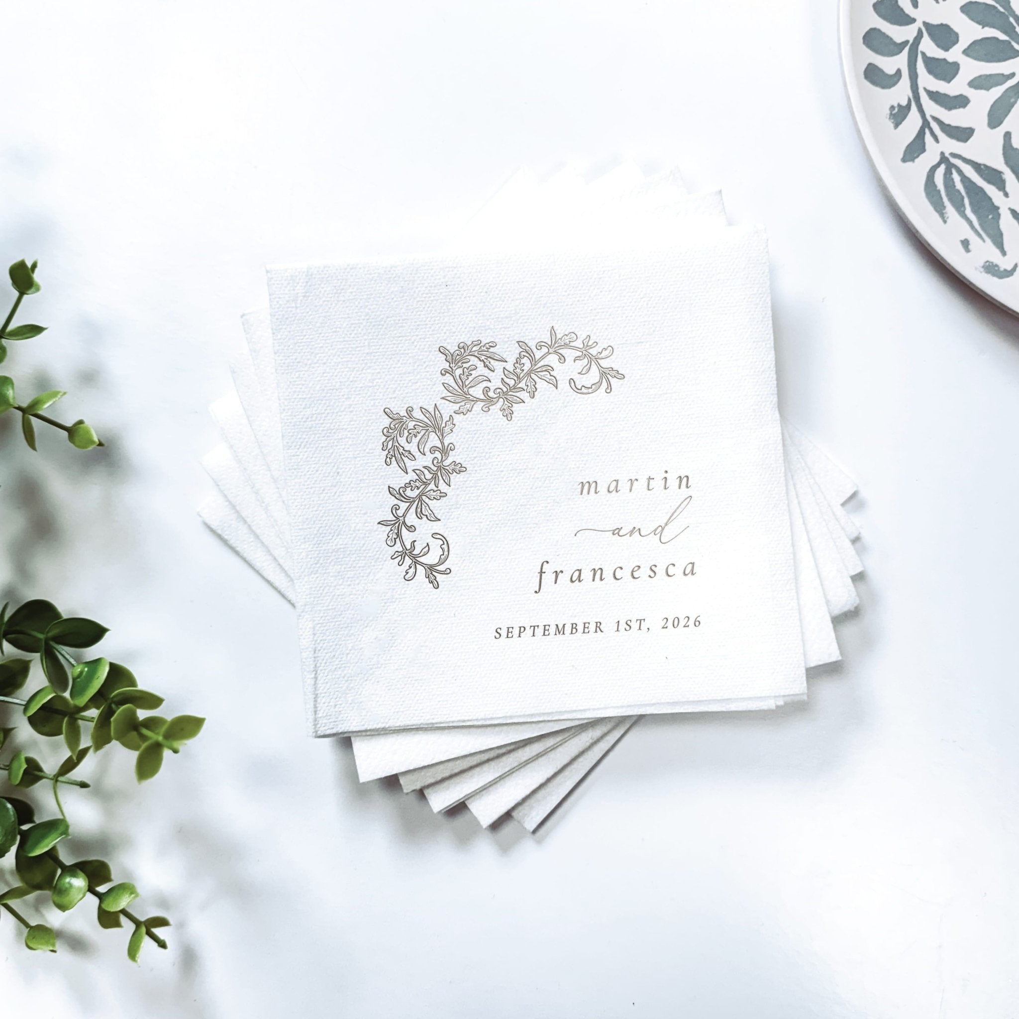 Stack of white cocktail napkins with delicate gray floral design and personalized names "marrin and francesca" with wedding date.