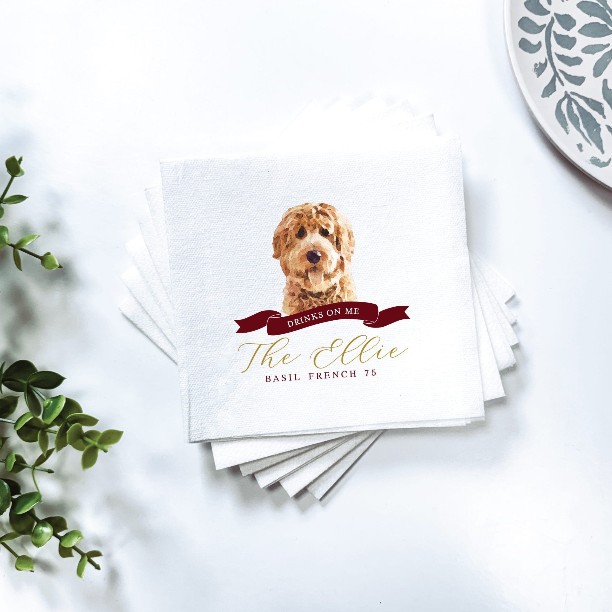 Stack of white cocktail napkins with golden doodle illustration, red ribbon banner, and "The Ollie" text personalization.