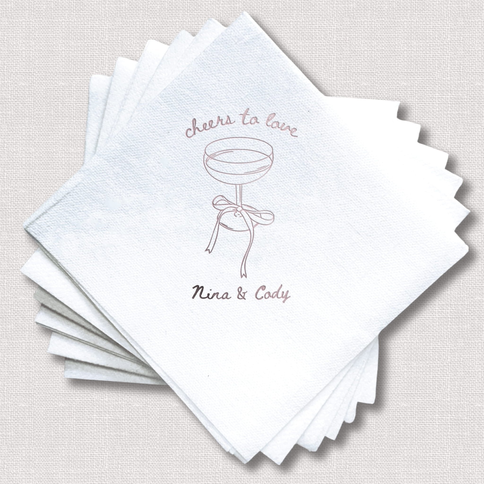 Stack of white linen napkins with "cheers to love" text and martini glass illustration, personalized with "Nina & Cody" in foil stamping.