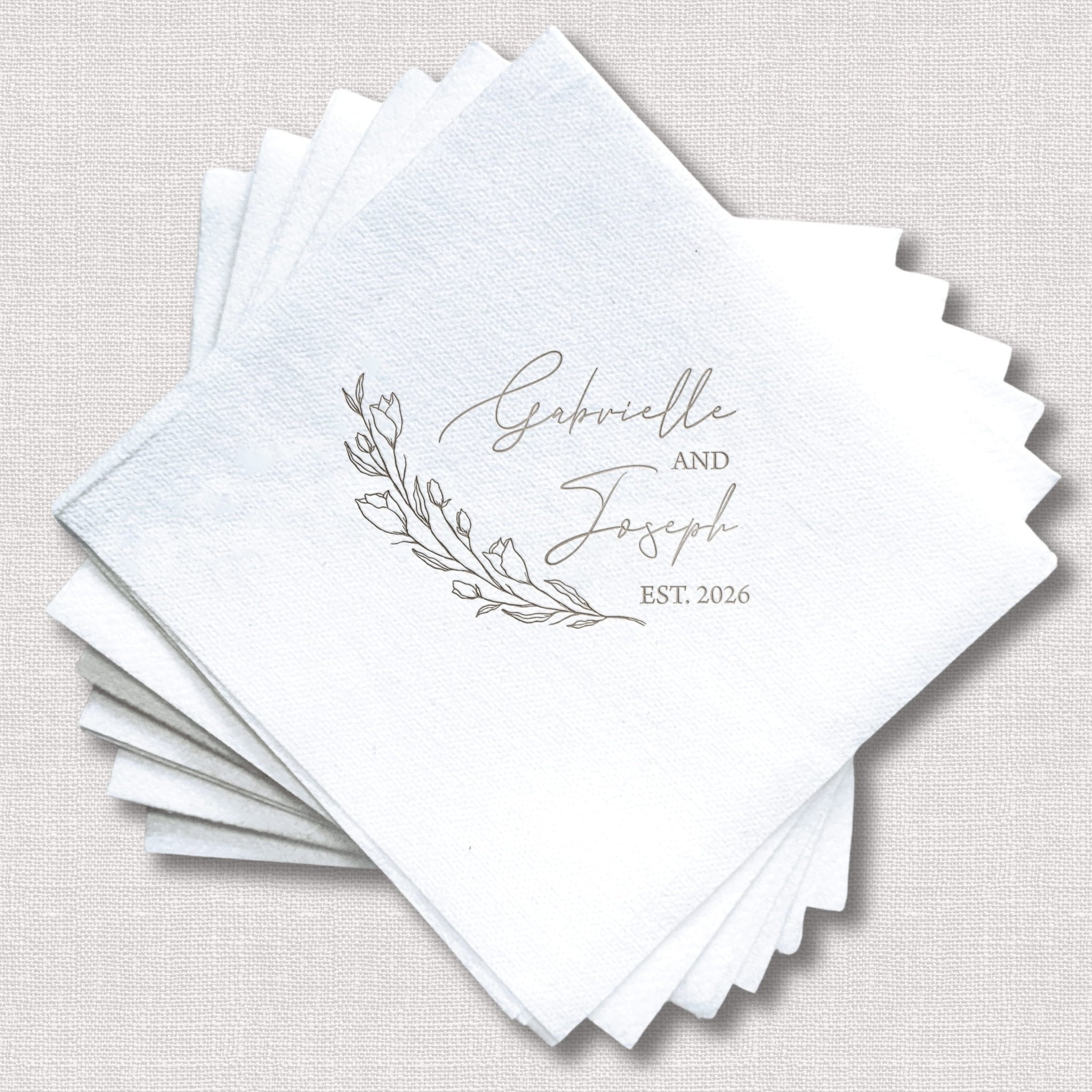 Stack of white linen napkins with "Gabriella and Joseph EST 2026" in gray foil stamping and delicate botanical leaf design