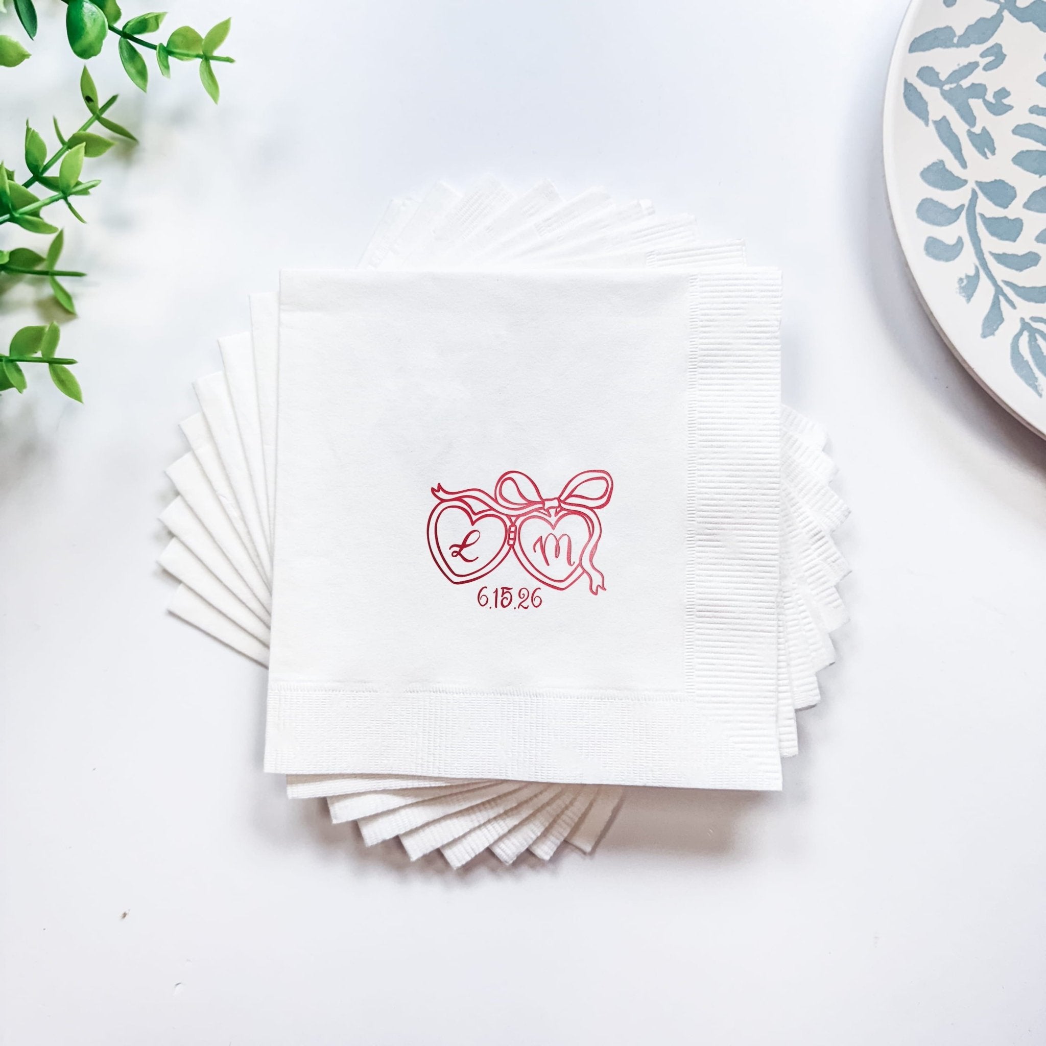 Stack of white cocktail napkins with pink foil-stamped heart locket design and initials "6.18.26"