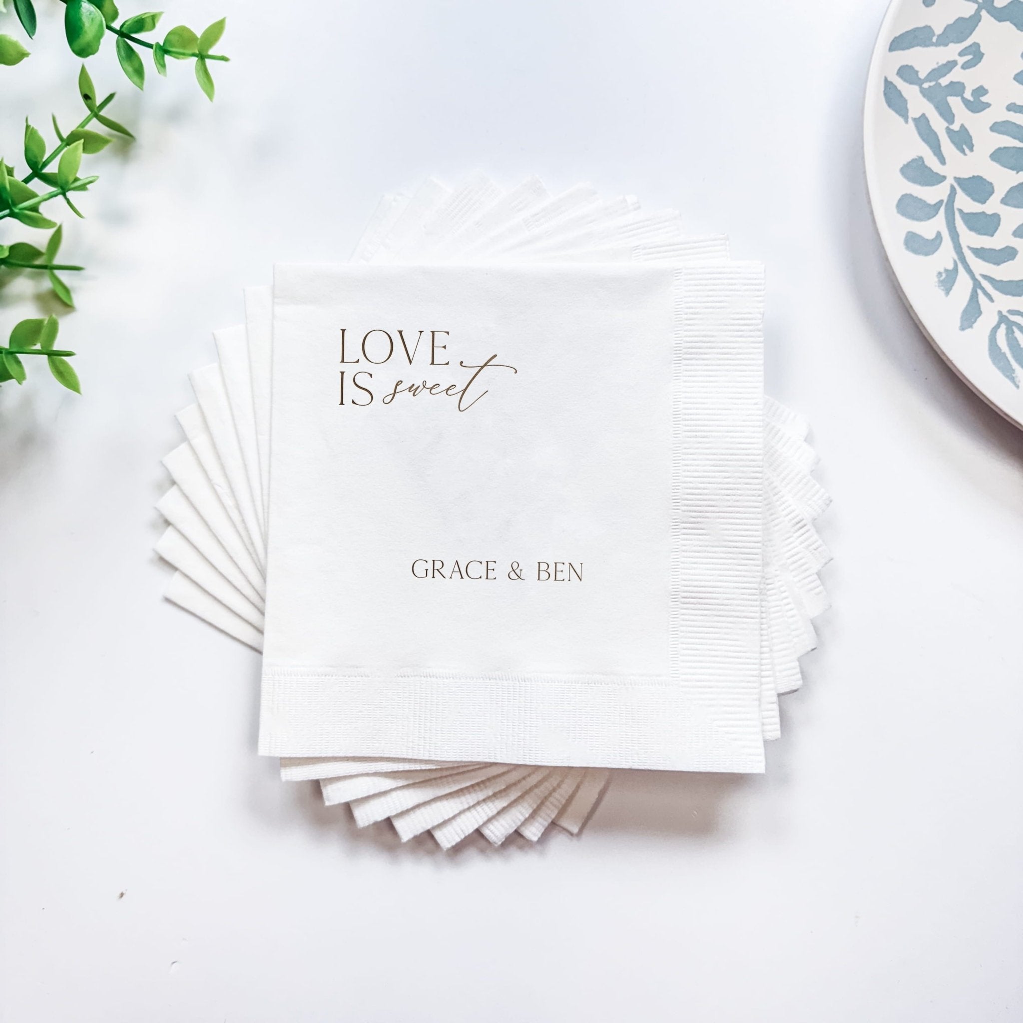 Stack of white foil-stamped napkins with "Love Is Sweet" text and personalized names "Grace & Ben" printed in gray