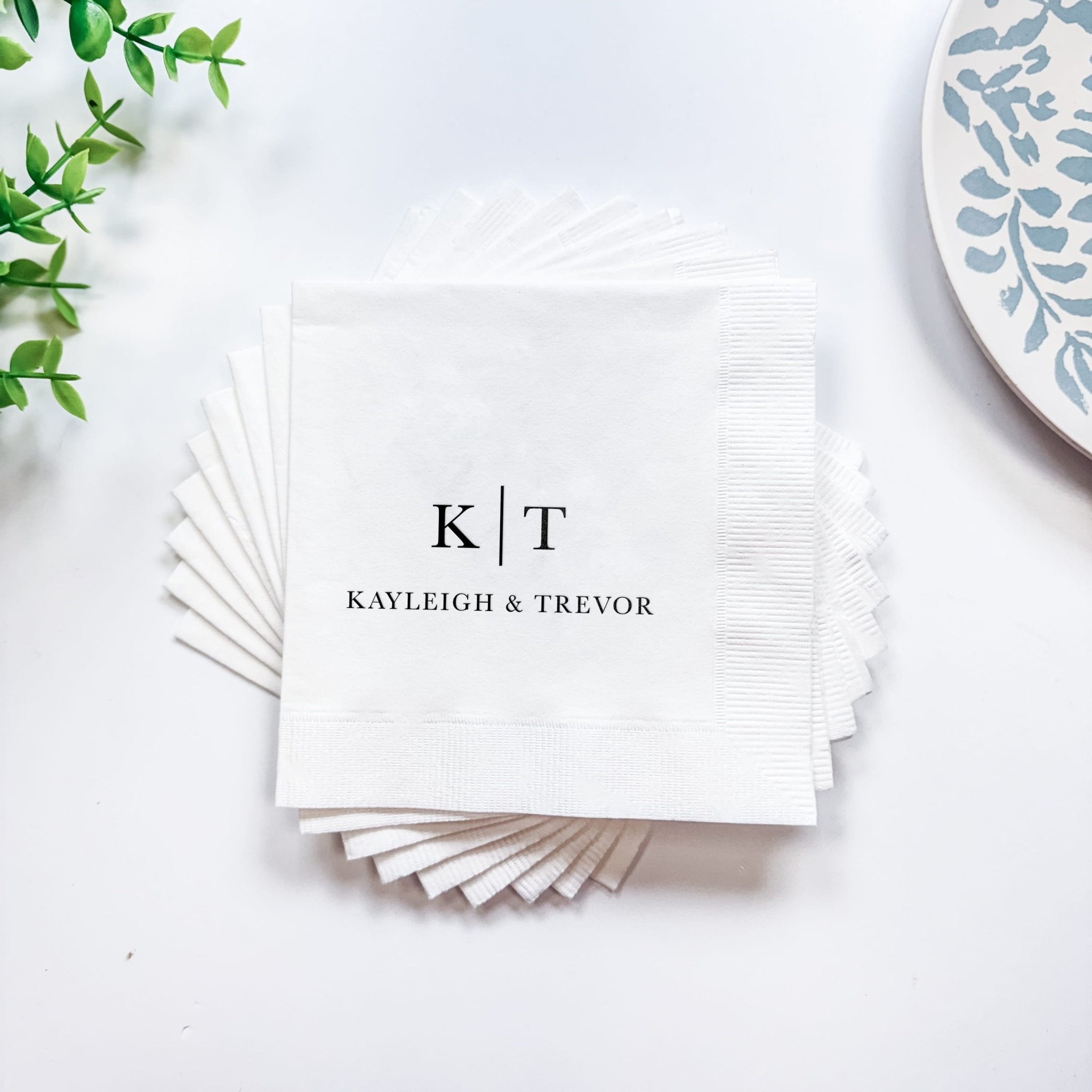 White cocktail napkins with initials "K|T" and names "KAYLEIGH & TREVOR" in modern foil-stamped design, fanned stack display.