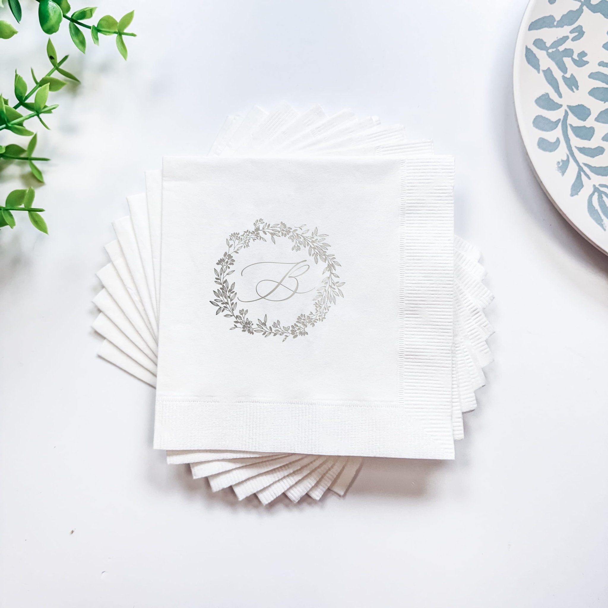 Stack of white napkins with silver foil stamped monogram wreath design and letter initial.