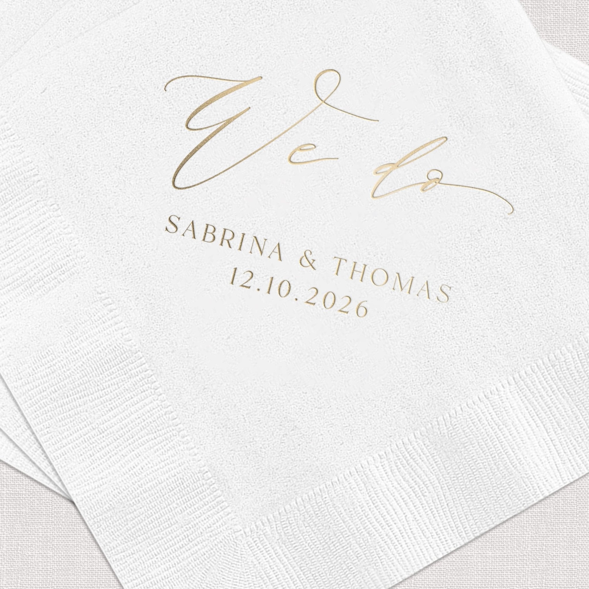 White napkins with gold foil "We Do" text and couple names Sabrina & Thomas with wedding date 12.10.2026