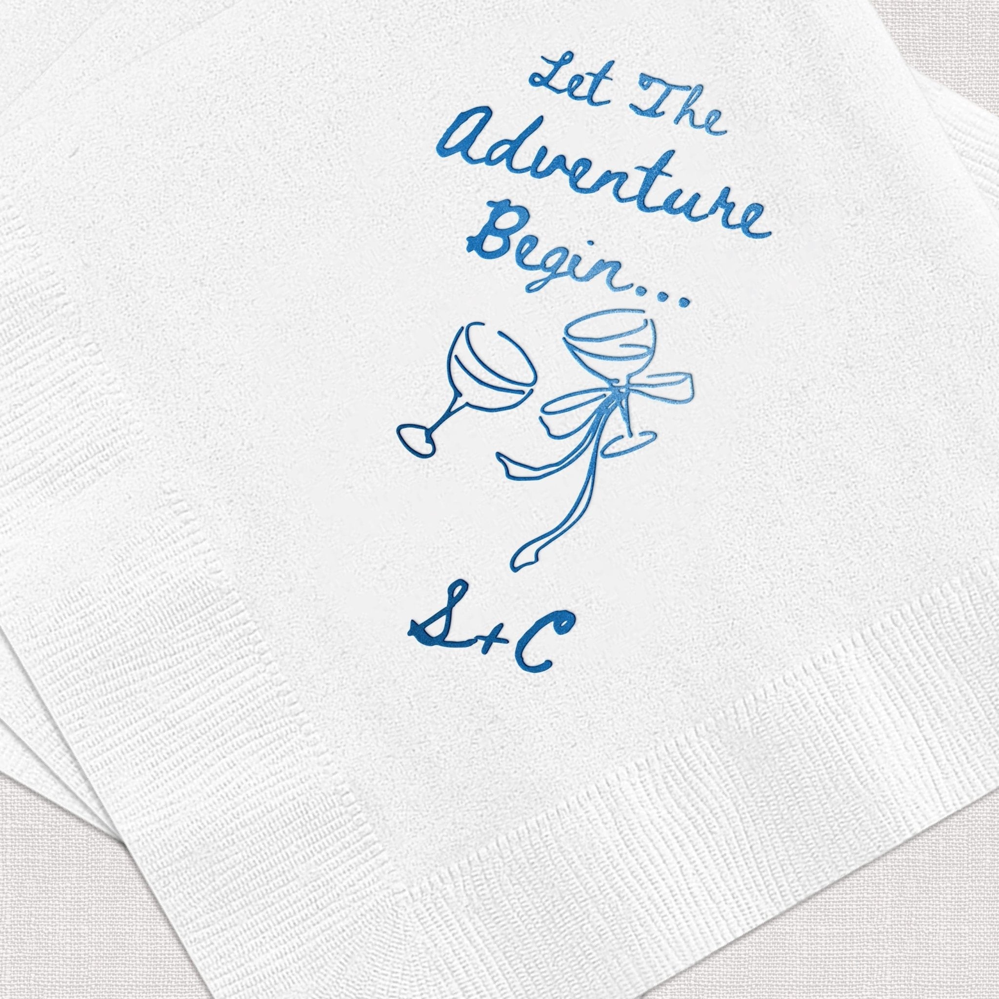 White cocktail napkins with blue foil stamped design featuring "Let The Adventure Begin" text, two champagne glasses, and initials "L&C"
