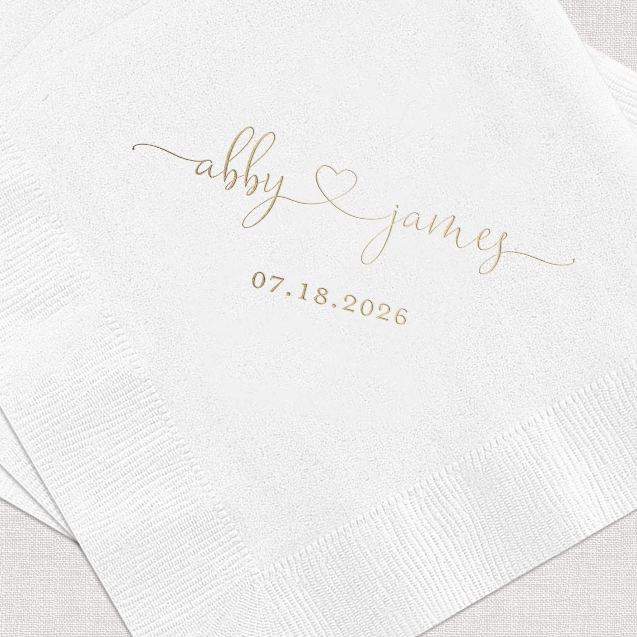 White cocktail napkins with gold foil stamped names "abby" and "james" with heart, and wedding date 07.18.2026
