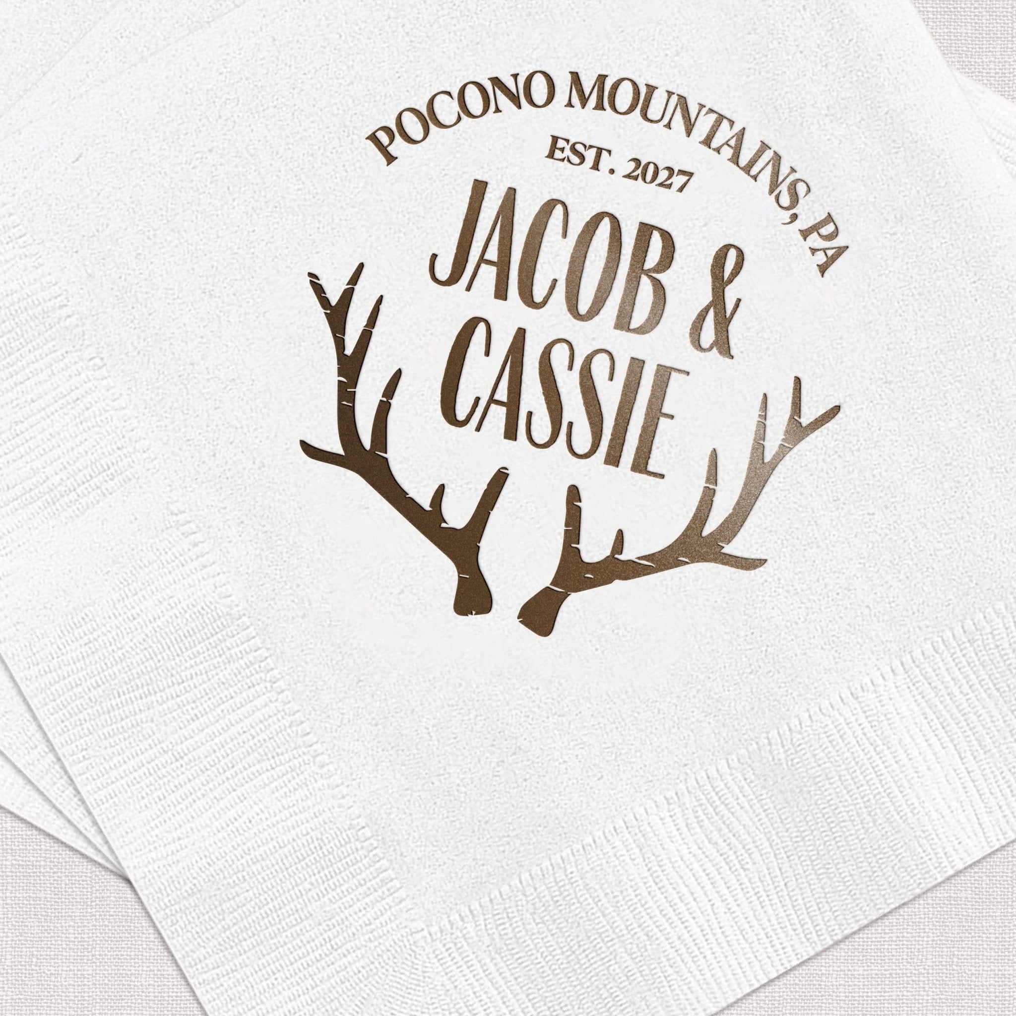 White napkin with brown foil-stamped antlers and personalized text "Jacob & Cassie" with "Pocono Mountains Est. 2027"