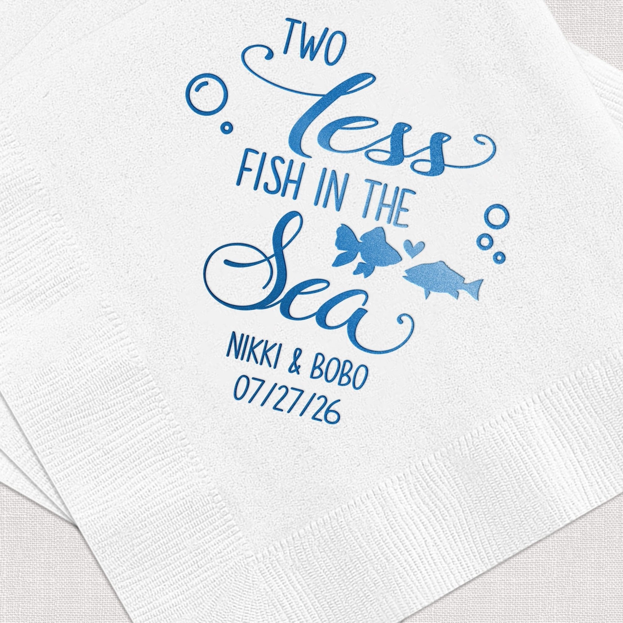 White cocktail napkin with blue foil-stamped design reading "Two Less Fish in the Sea" with fish graphics, personalized names and wedding date.