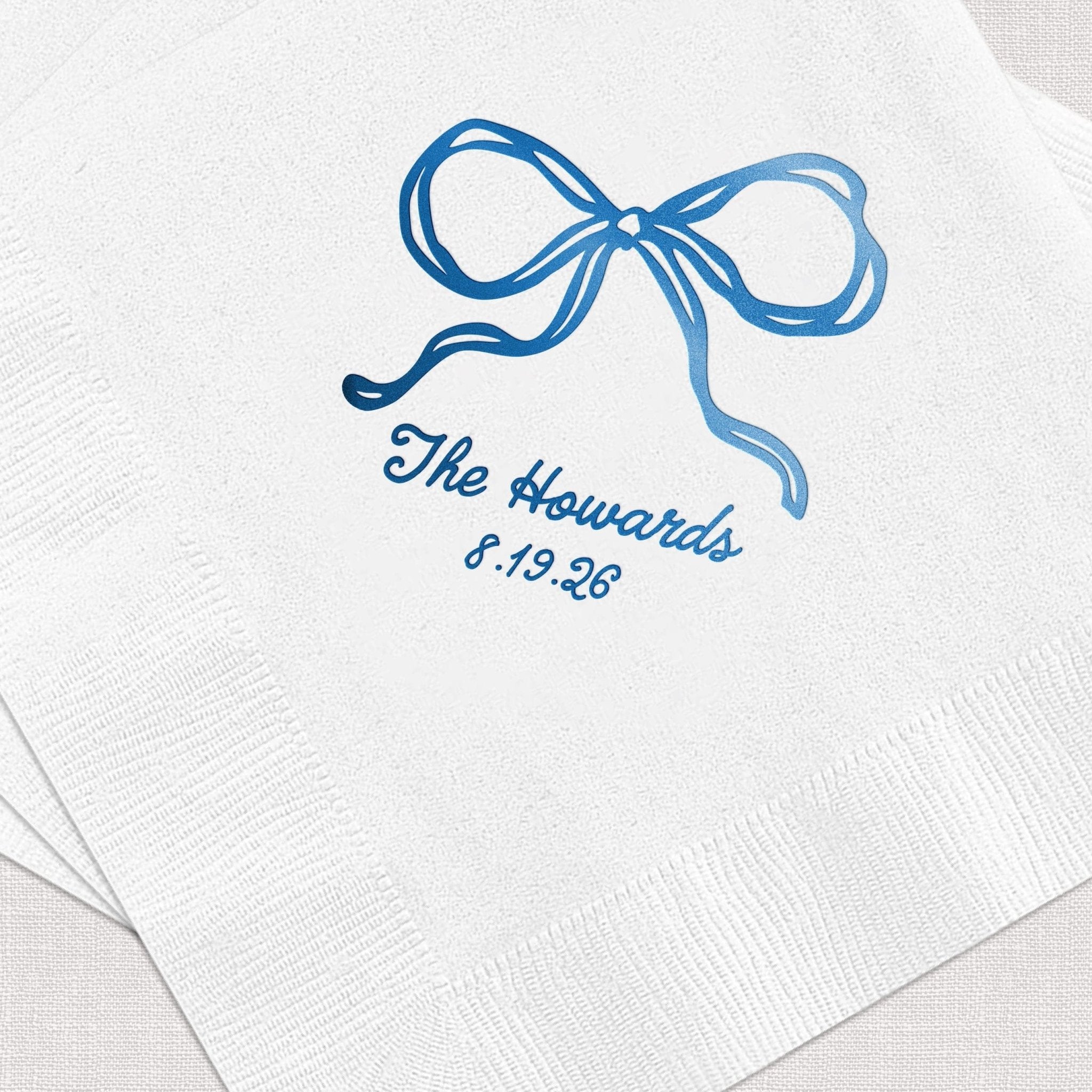 White cocktail napkins with blue foil stamped bow design and personalized text reading "The Howards 5.19.25"