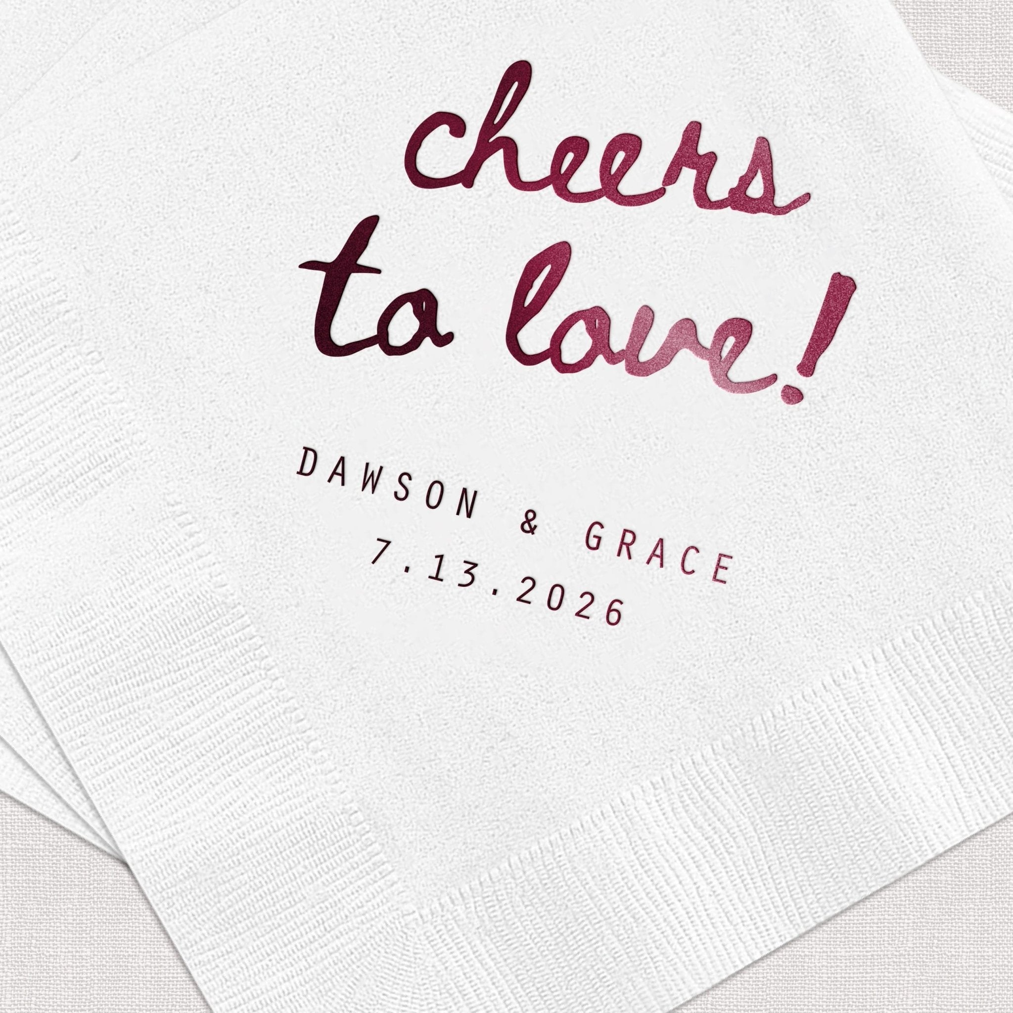 White napkin with burgundy foil-stamped text reading "cheers to love!" and personalized names "Dawson & Grace" with wedding date 7.13.2026