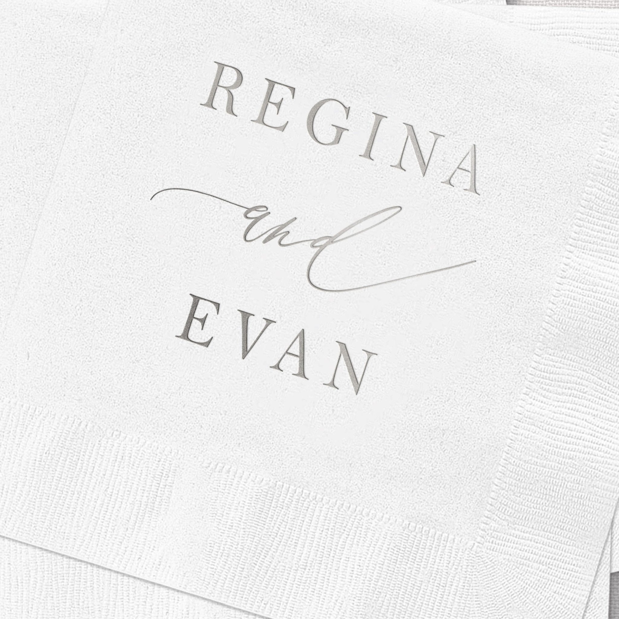 White cocktail napkins with silver foil stamped text reading "REGINA and EVAN" with decorative script styling