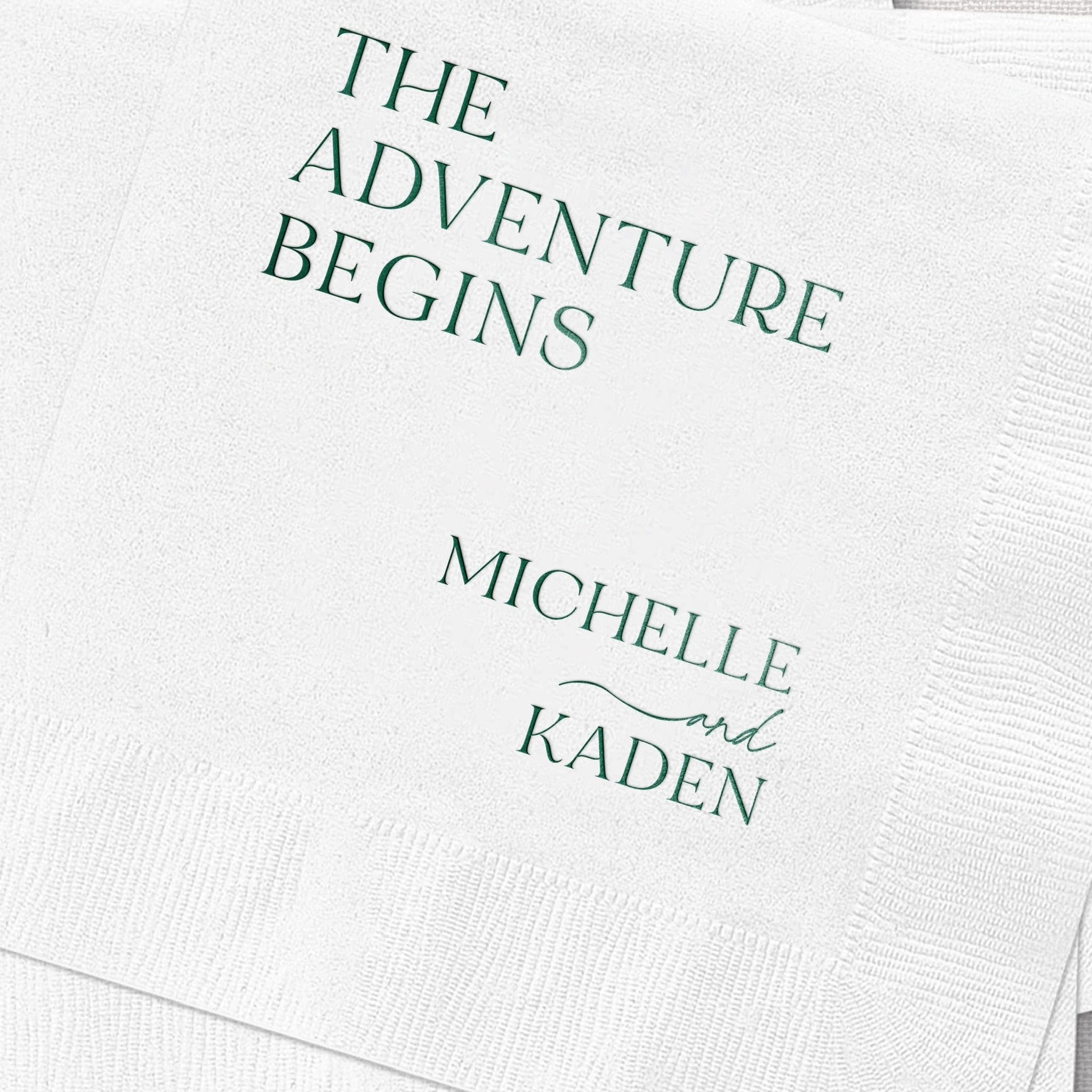 White cocktail napkins with teal foil stamping reading "The Adventure Begins" and personalized names "Michelle and Kaden"