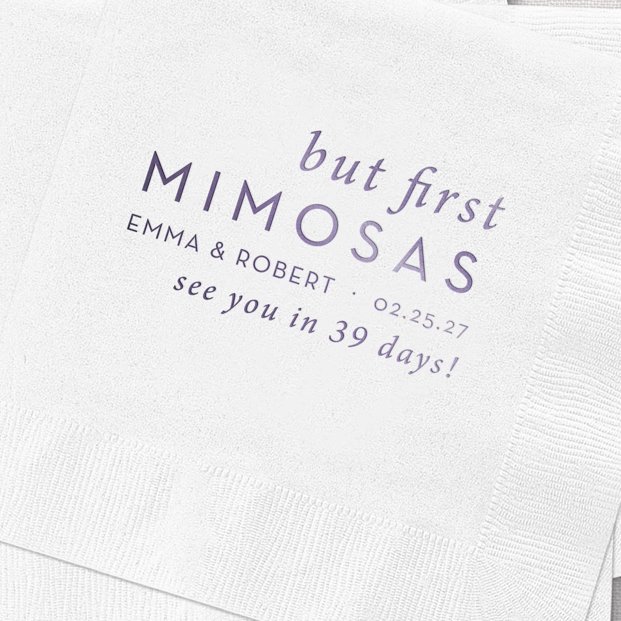 White cocktail napkin with navy foil-stamped text reading "but first MIMOSAS" and personalization with names, date, and "see you in 39 days!"
