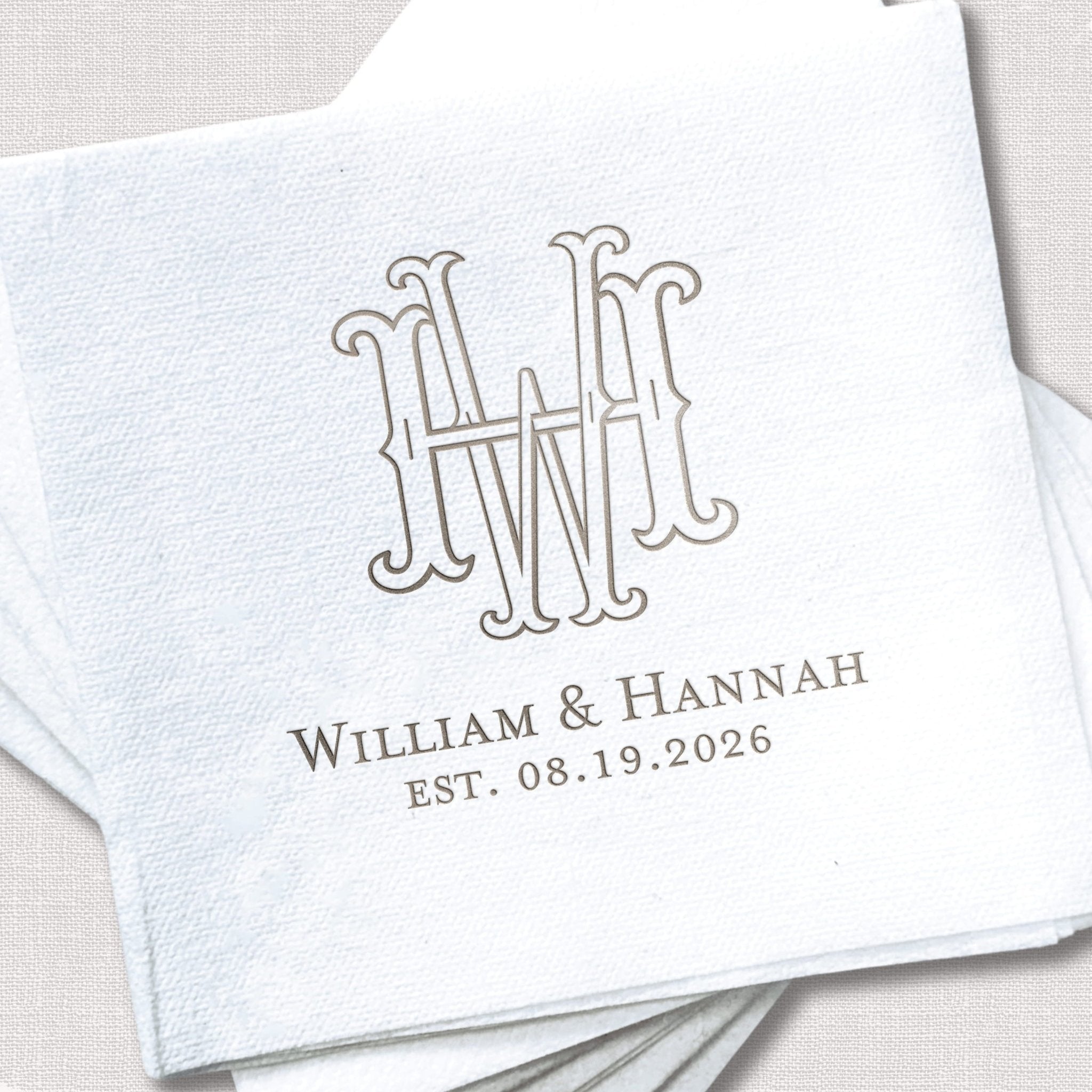 White linen napkin with interlocking monogram and "William & Hannah EST. 08.19.2026" in gray foil stamping.