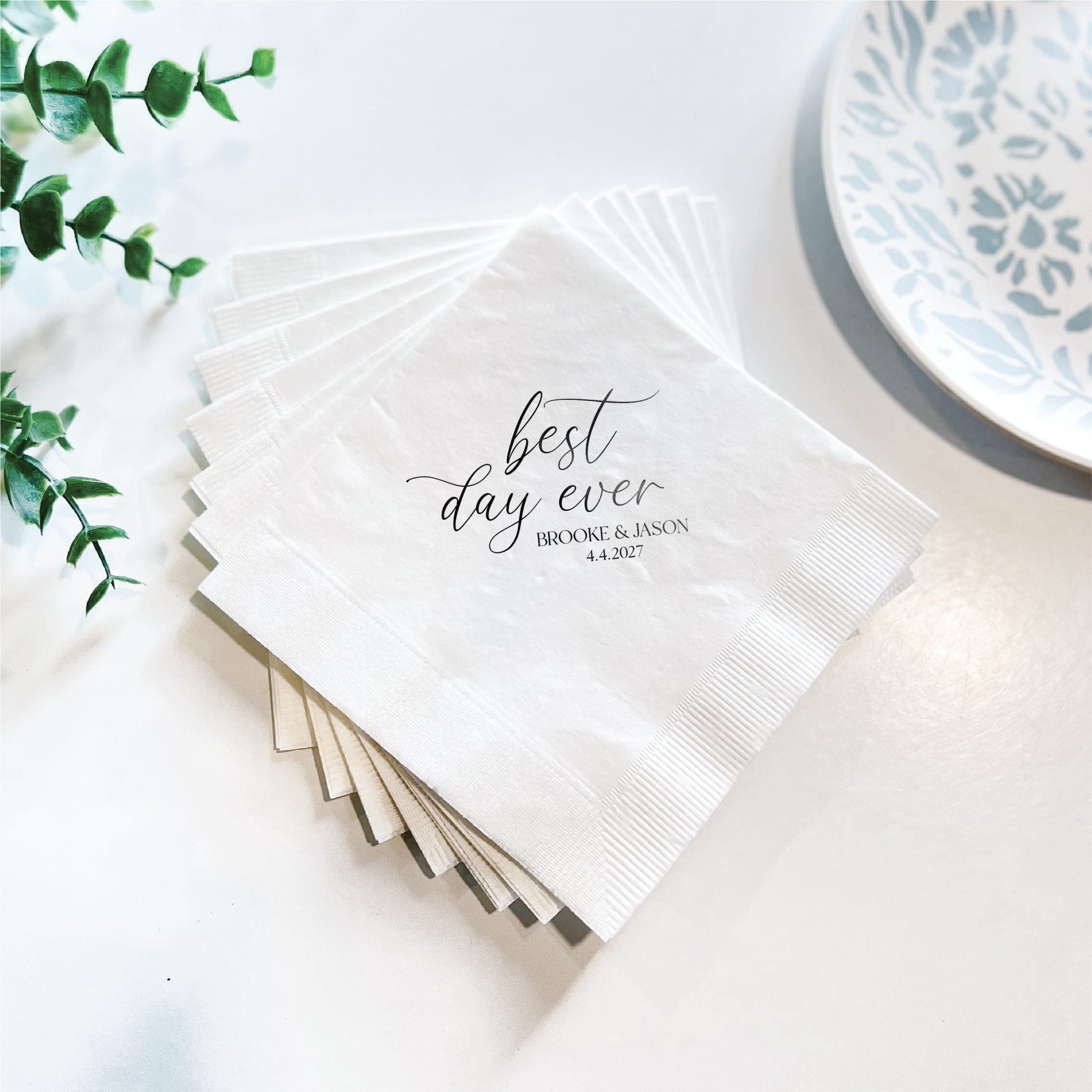 Stack of white napkins with "best day ever" script text and personalized names foil stamped in gray, styled with greenery and blue patterned plate.