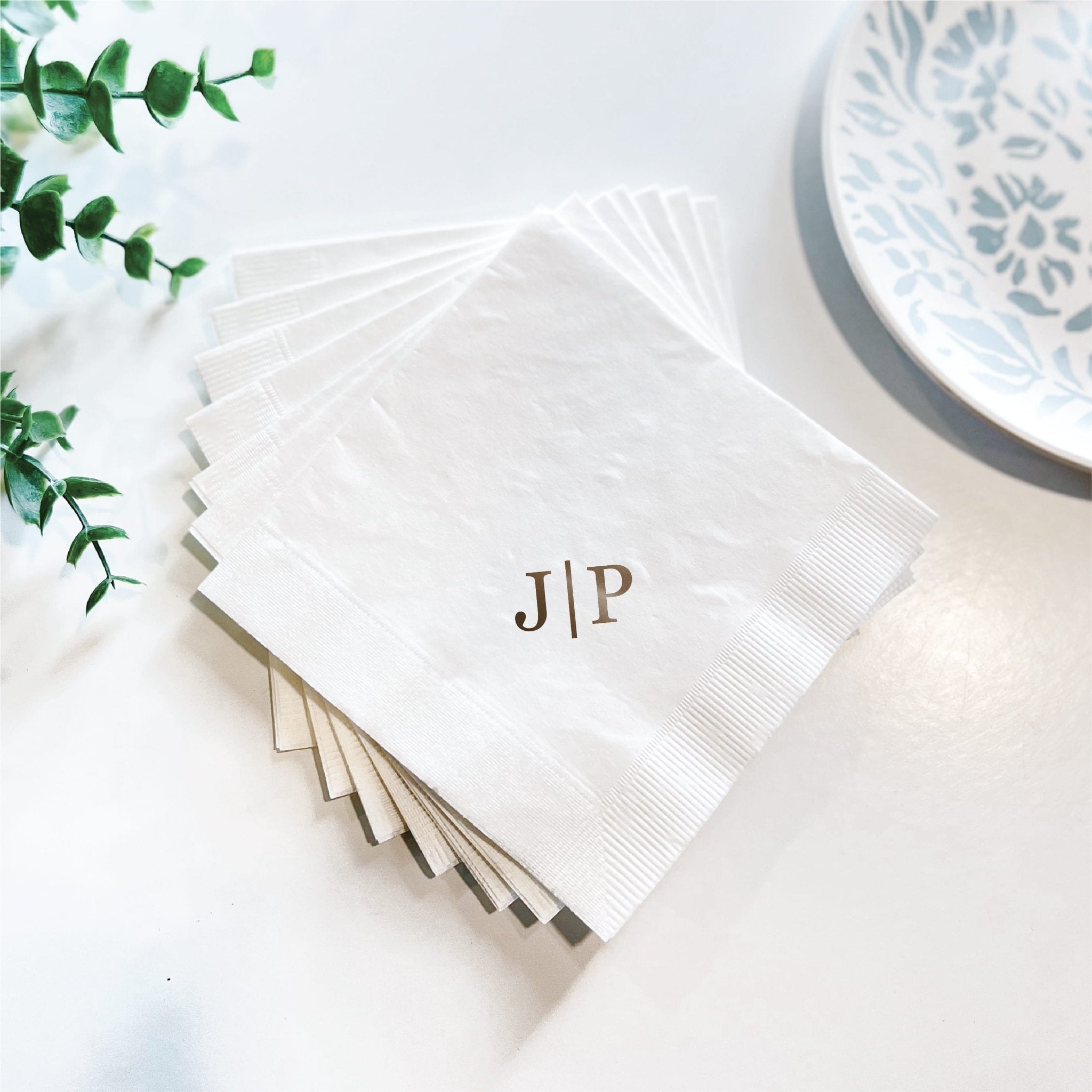 Stack of white cocktail napkins with gold monogram initials JIP, displayed with eucalyptus and blue patterned plate