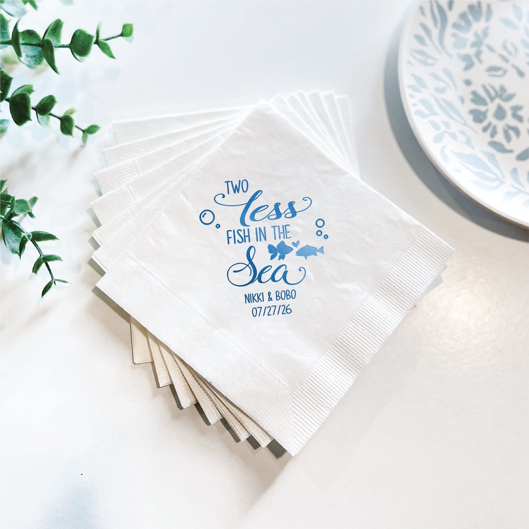 Stack of white cocktail napkins with blue foil-stamped design reading "Two Less Fish in the Sea" and personalized names and date.