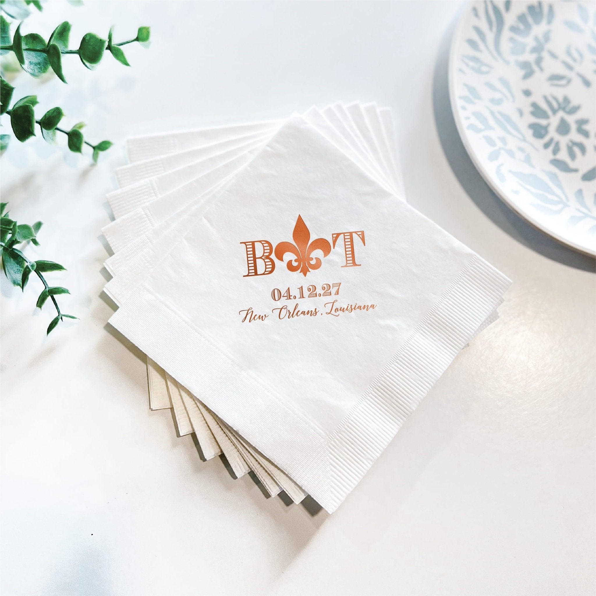 Stack of white cocktail napkins with orange foil-stamped fleur de lis design and personalized text reading "BOAT 04.13.27 Two Fabulous Grooms"