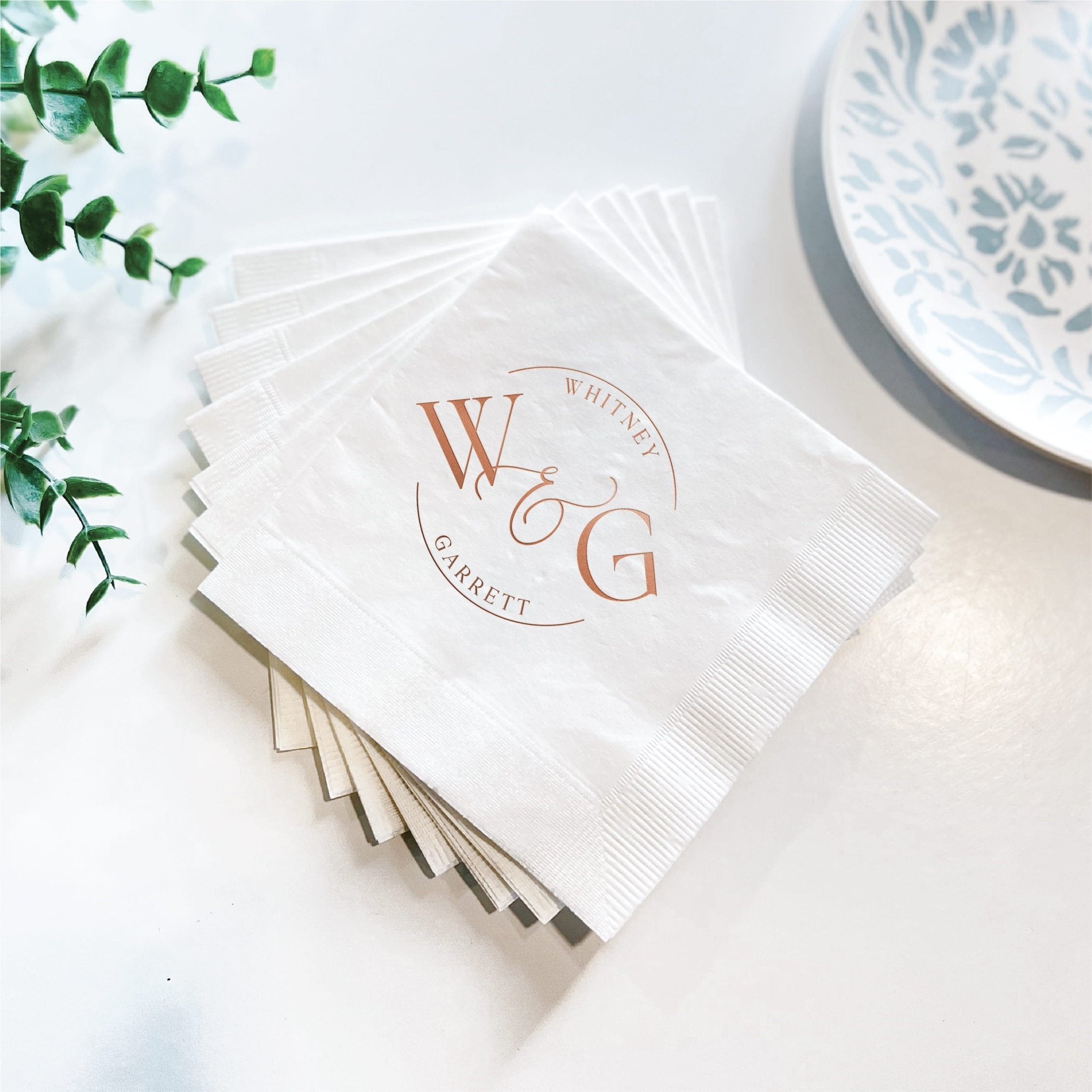Stack of white napkins with rose gold foil stamped circular monogram design featuring initials WG and wedding date text.