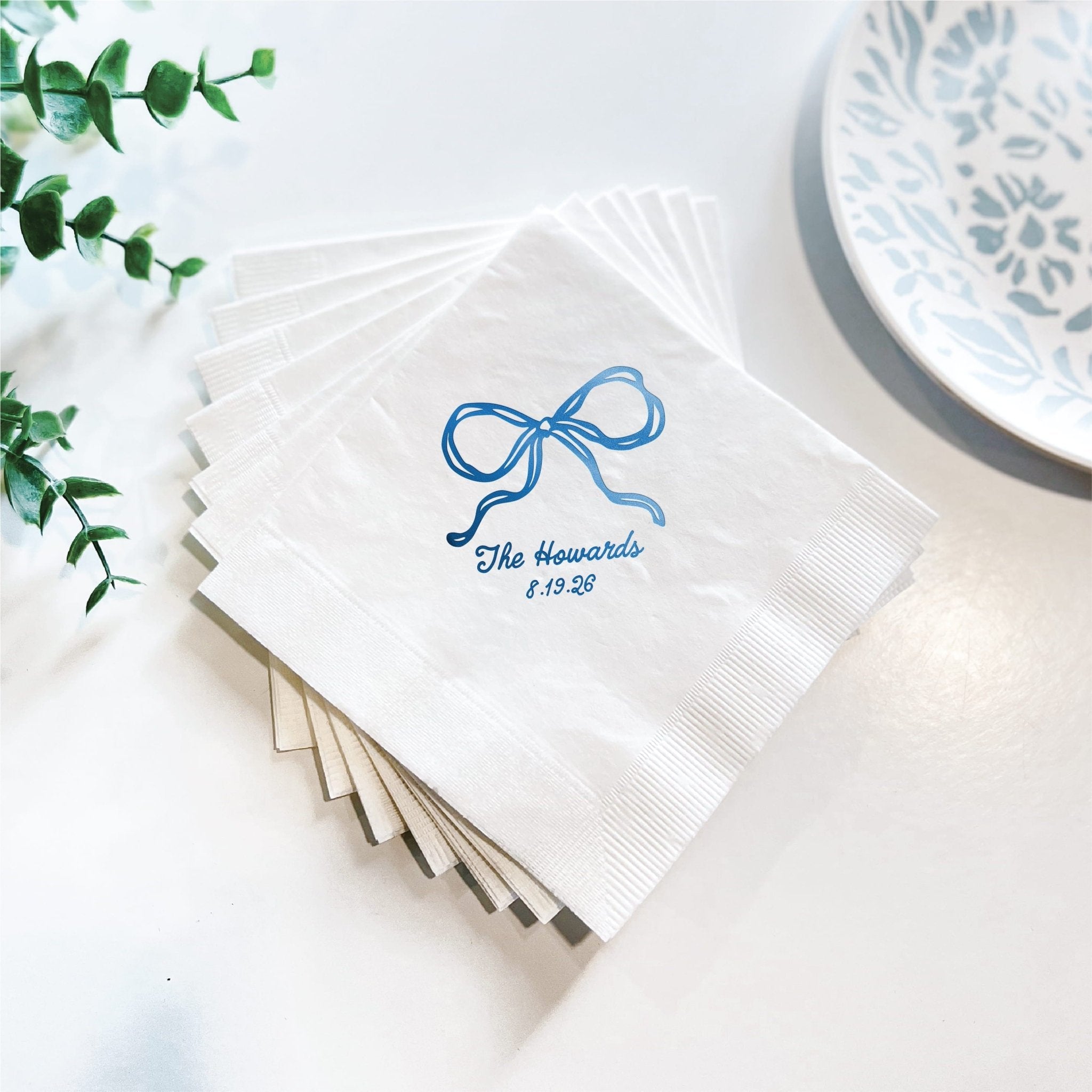Stack of white cocktail napkins with blue foil-stamped bow design and "The Howells 4.15.23" personalization text