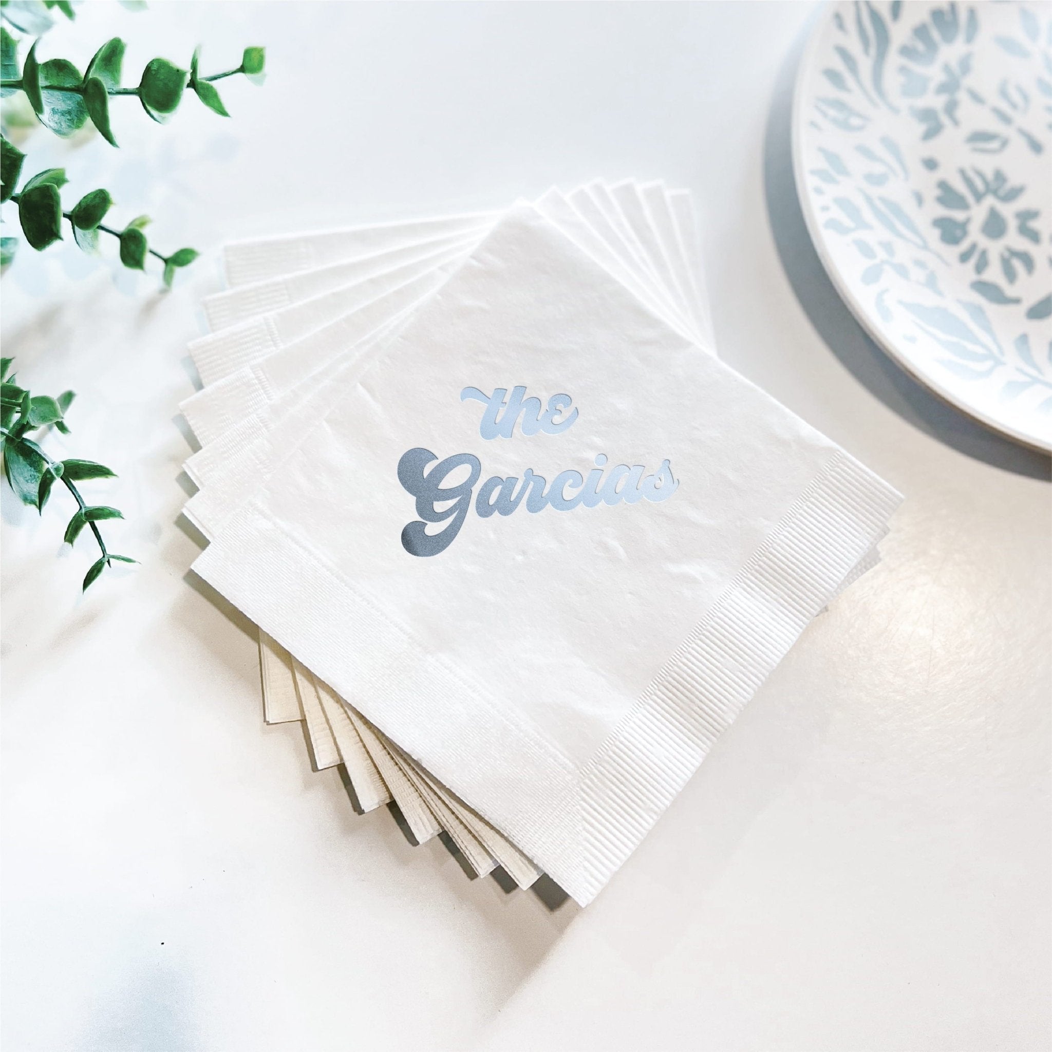 Stack of white cocktail napkins with blue foil-stamped "the Garcias" text, displayed with greenery and coordinating plate