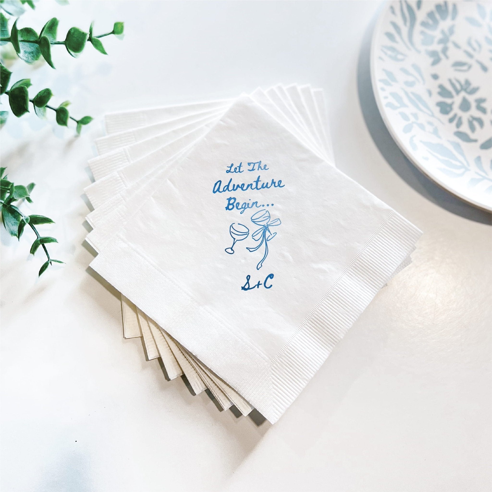 Stack of white napkins with blue foil stamped text reading "Let The Adventure Begin" and couple illustration, styled with greenery and decorative plate.