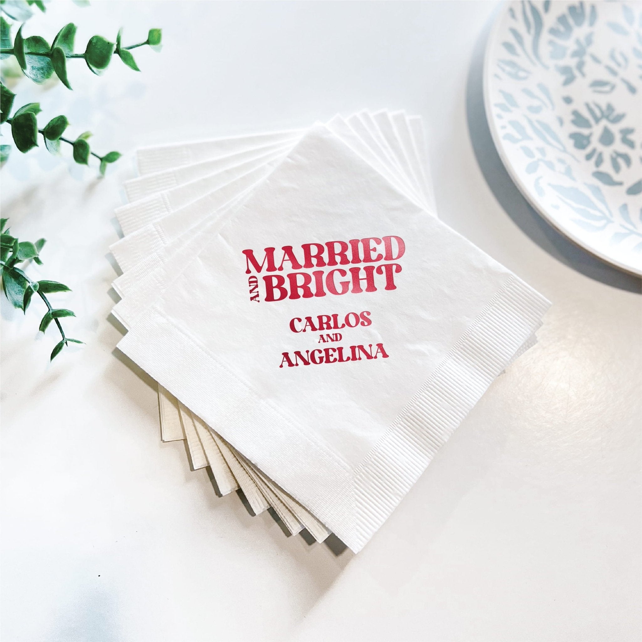 Stack of white napkins with red foil stamped text reading "Married and Bright" and couple's names Carlos and Angelina