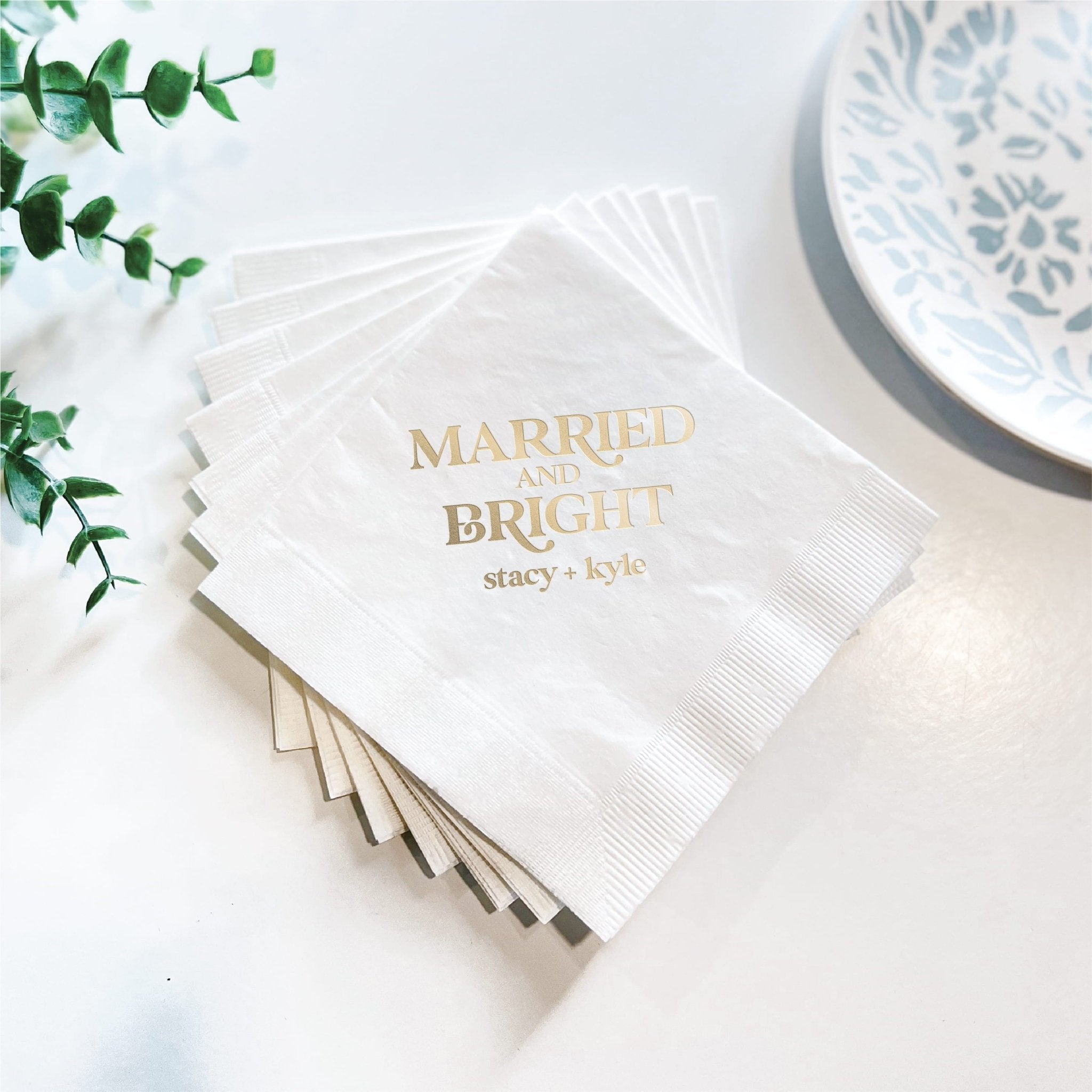Stack of white napkins with gold foil "Married and Bright" text and personalized names, displayed with greenery and decorative plate.