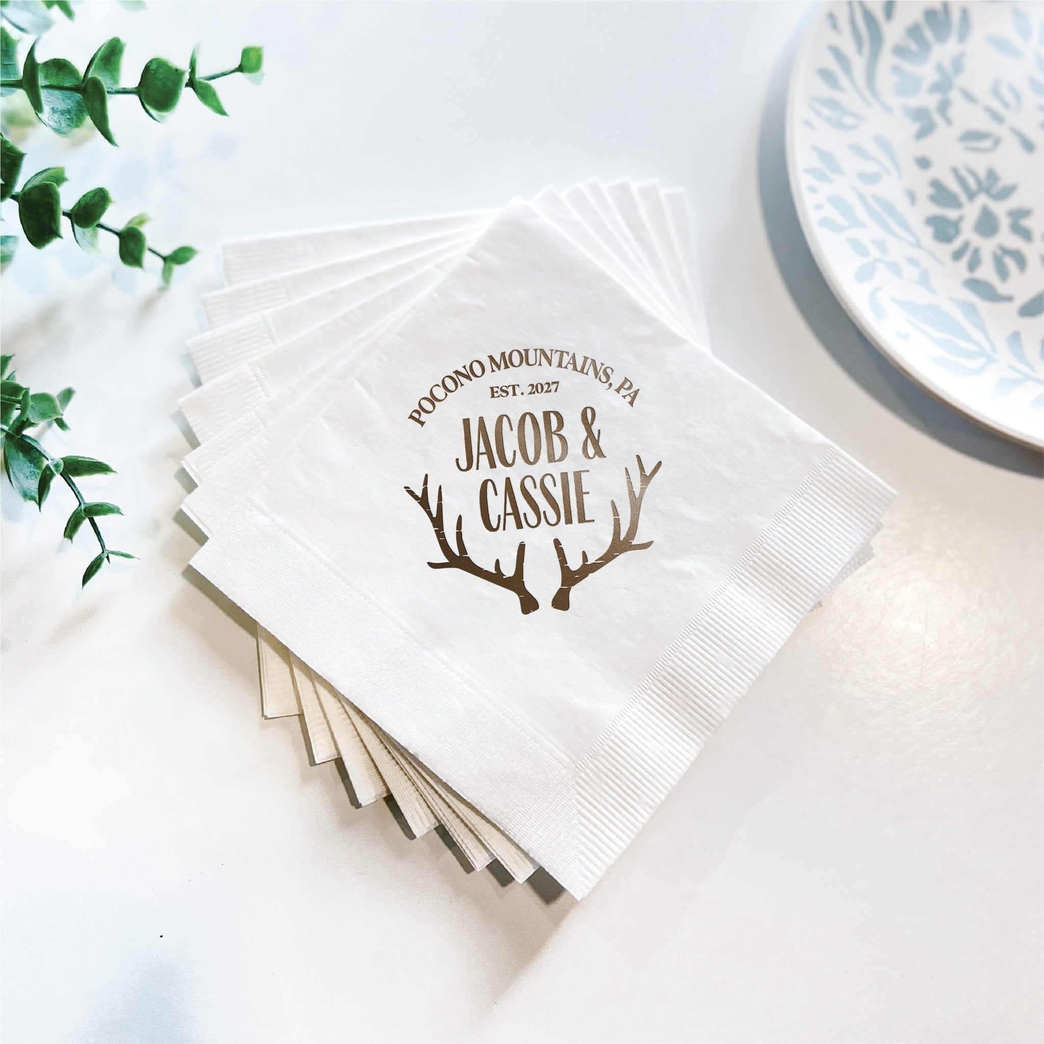 Stack of white personalized napkins with brown antler design and "Jacob & Cassie" text with wedding date.
